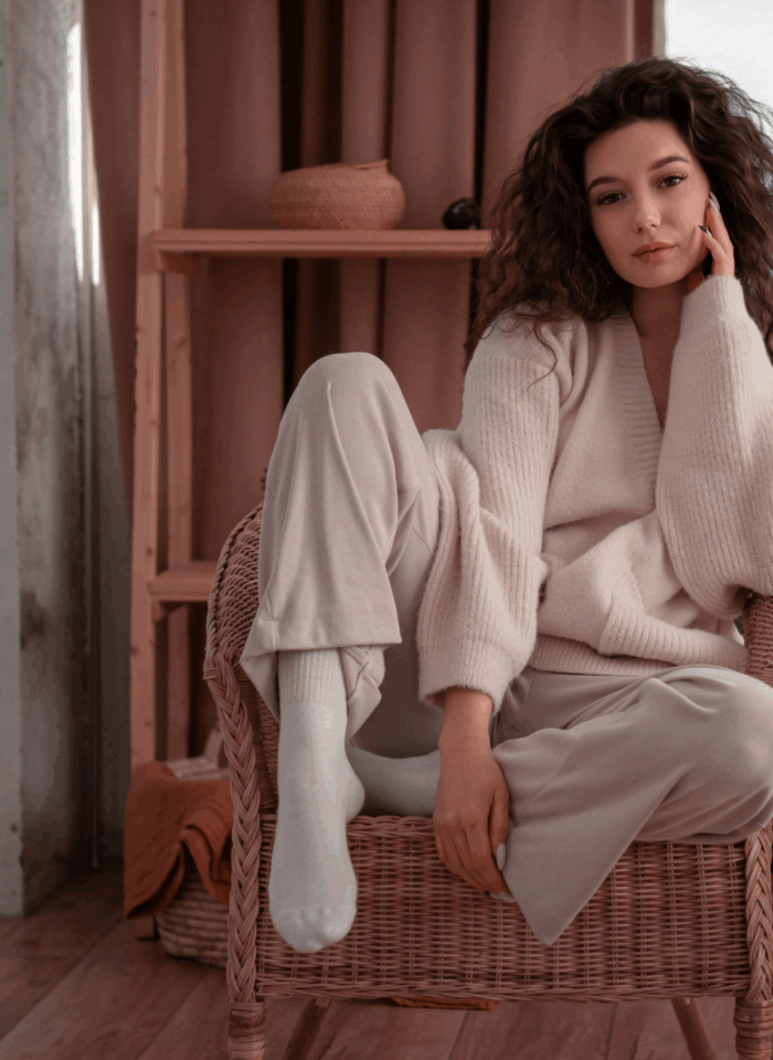 A woman dressed in cozy neutral loungewear sitting comfortably on a wicker chair in a warm and softly lit room.