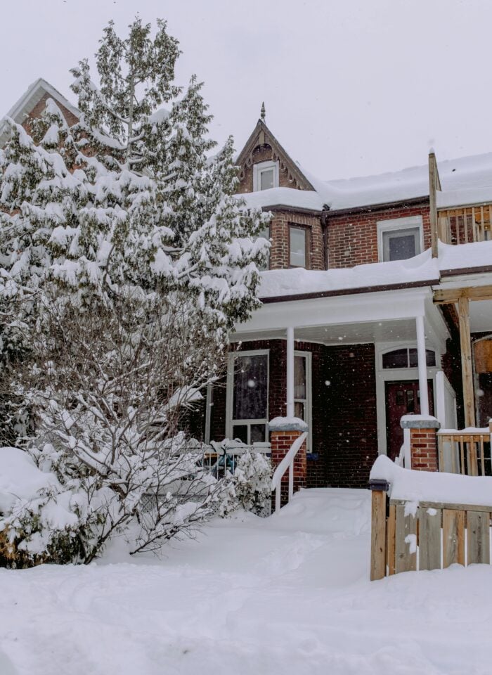 A brick house blanketed in snow, with a snow-laden front porch, roof, trees, and garden during a winter snowfall.