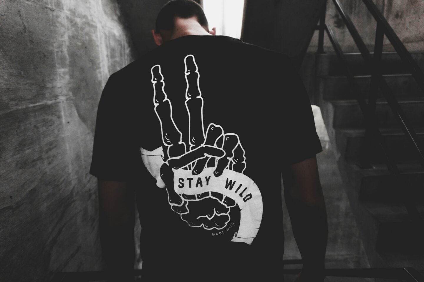 Back view of a person wearing a black t-shirt with a white graphic of a skeleton hand making a peace sign and the words "STAY WILD" printed across a ribbon.