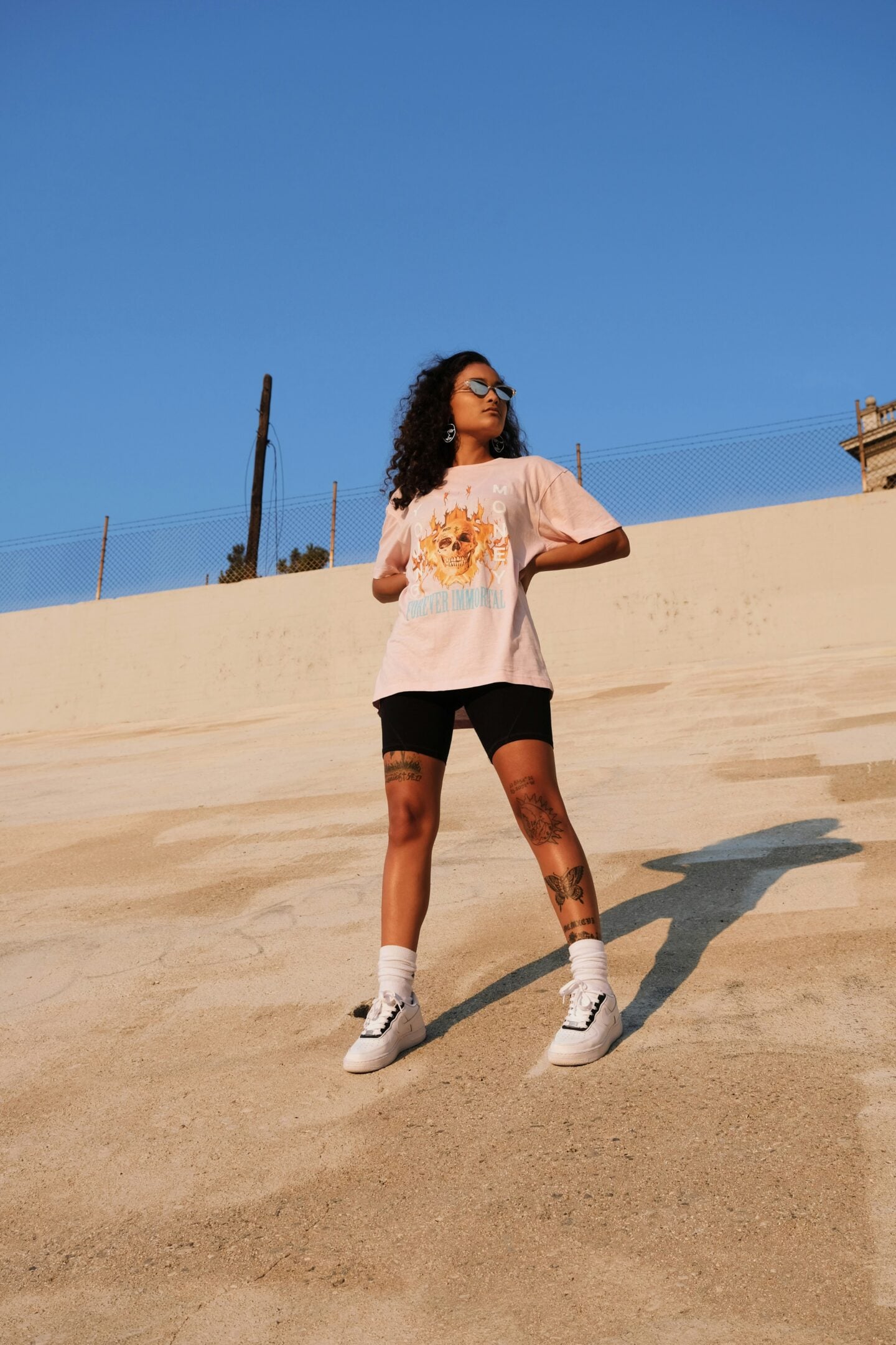 Stylish woman posing outdoors in a pastel pink graphic t-shirt that reads “FOREVER IMMORTAL,” paired with black biker shorts, sunglasses, and white sneakers.