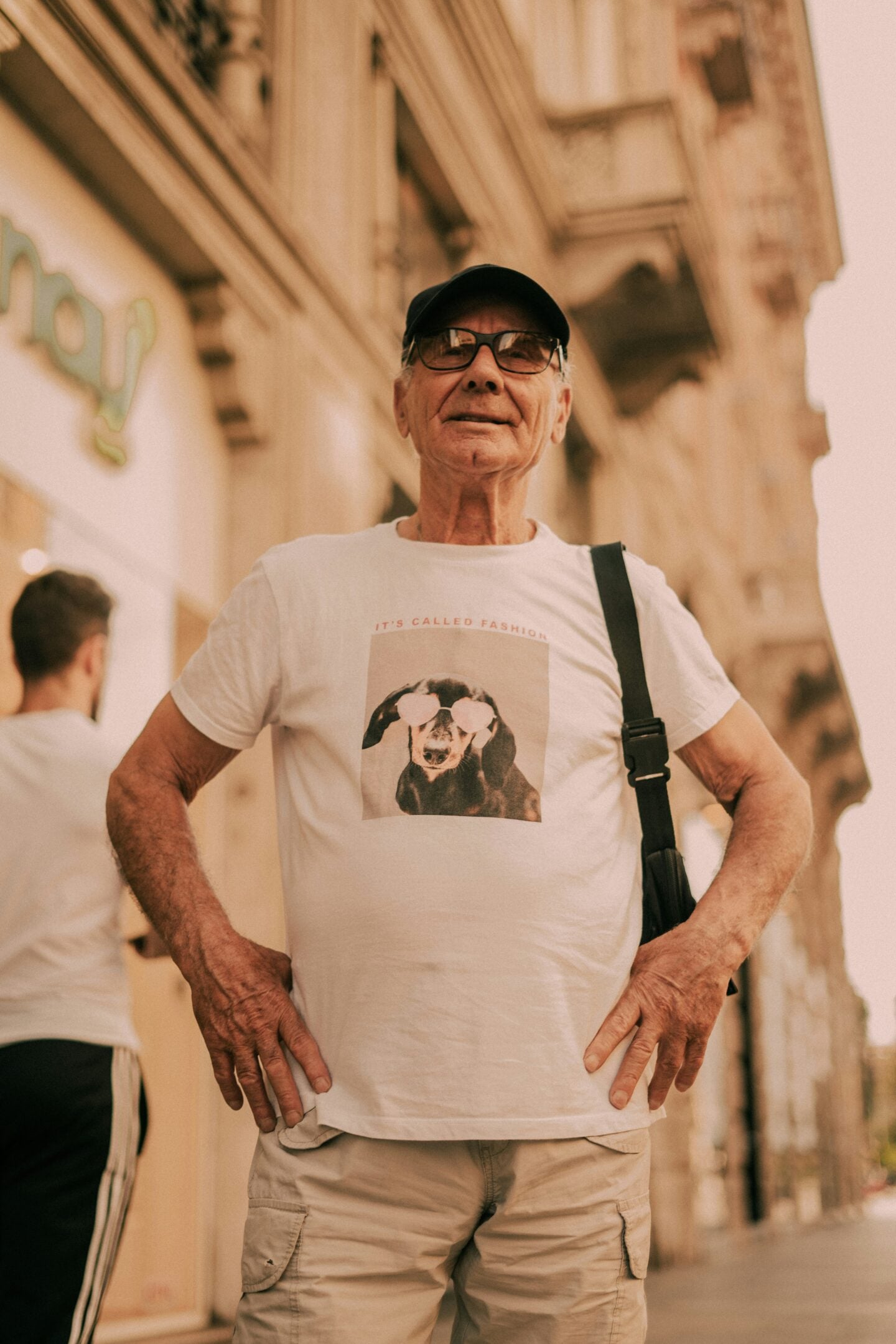 Elderly man smiling confidently in a white t-shirt with a photo of a dog wearing heart-shaped sunglasses and the caption "IT'S CALLED FASHION."
