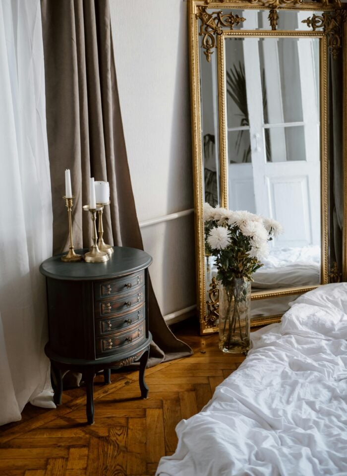 Elegant bedroom corner with a vintage black nightstand, brass candlesticks, a large ornate gold mirror, and a vase of white chrysanthemums beside an unmade bed with white sheets.