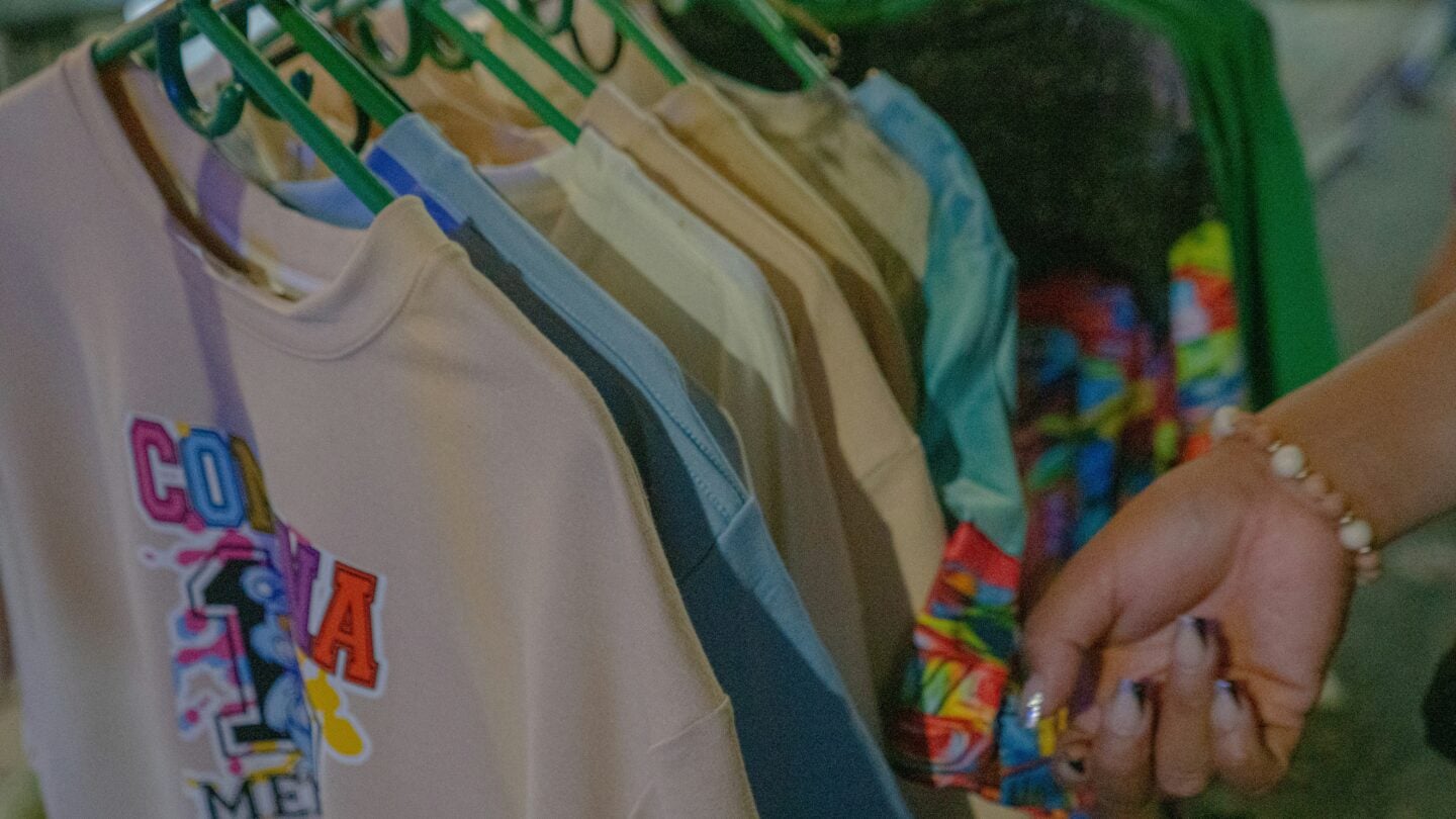 Close-up of a hand browsing through colorful graphic t-shirts on green hangers at a clothing rack, with one shirt prominently featuring cartoon-style lettering.