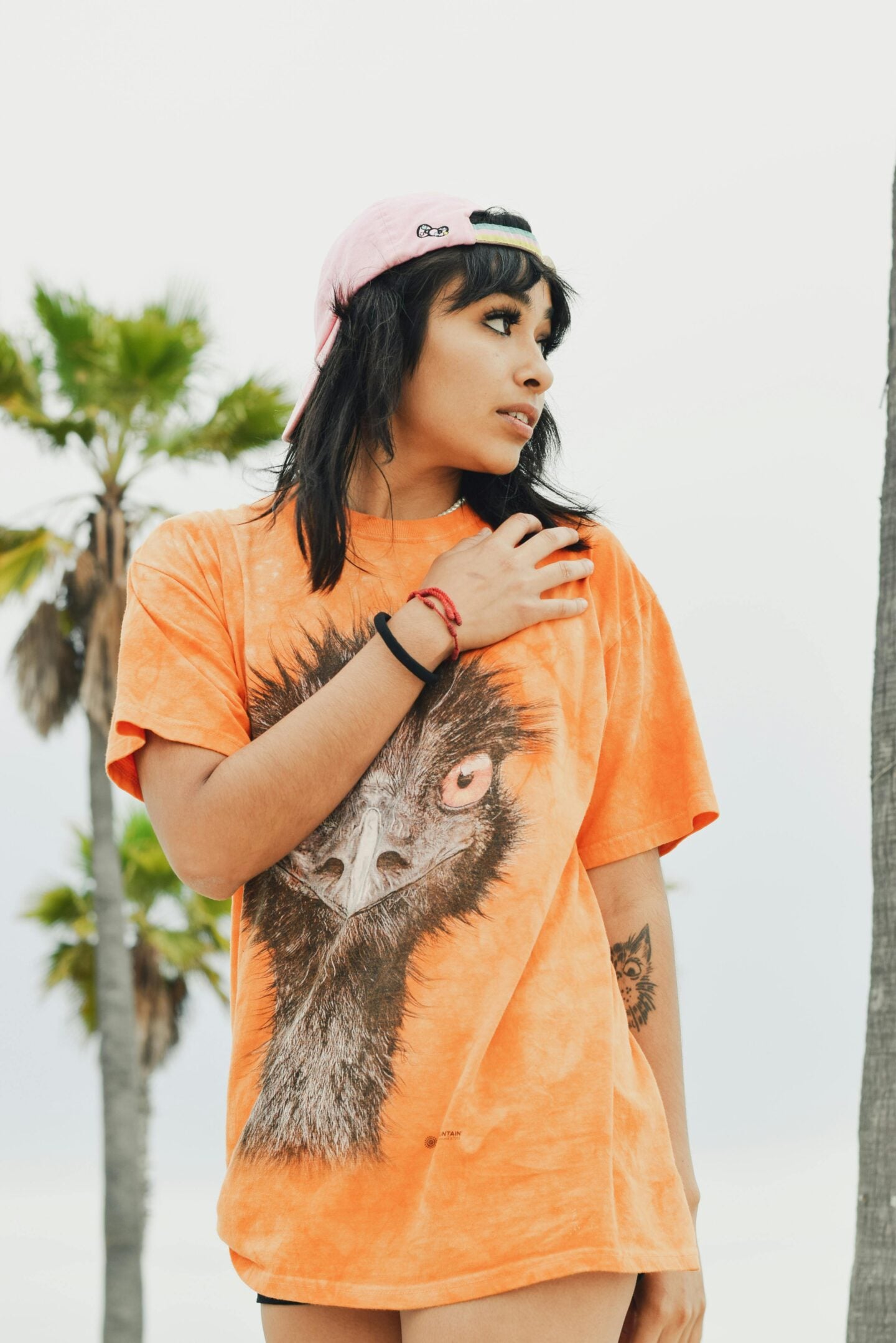 Young woman in an orange t-shirt with a realistic ostrich face print, wearing a backward pink cap and standing against a backdrop of palm trees.