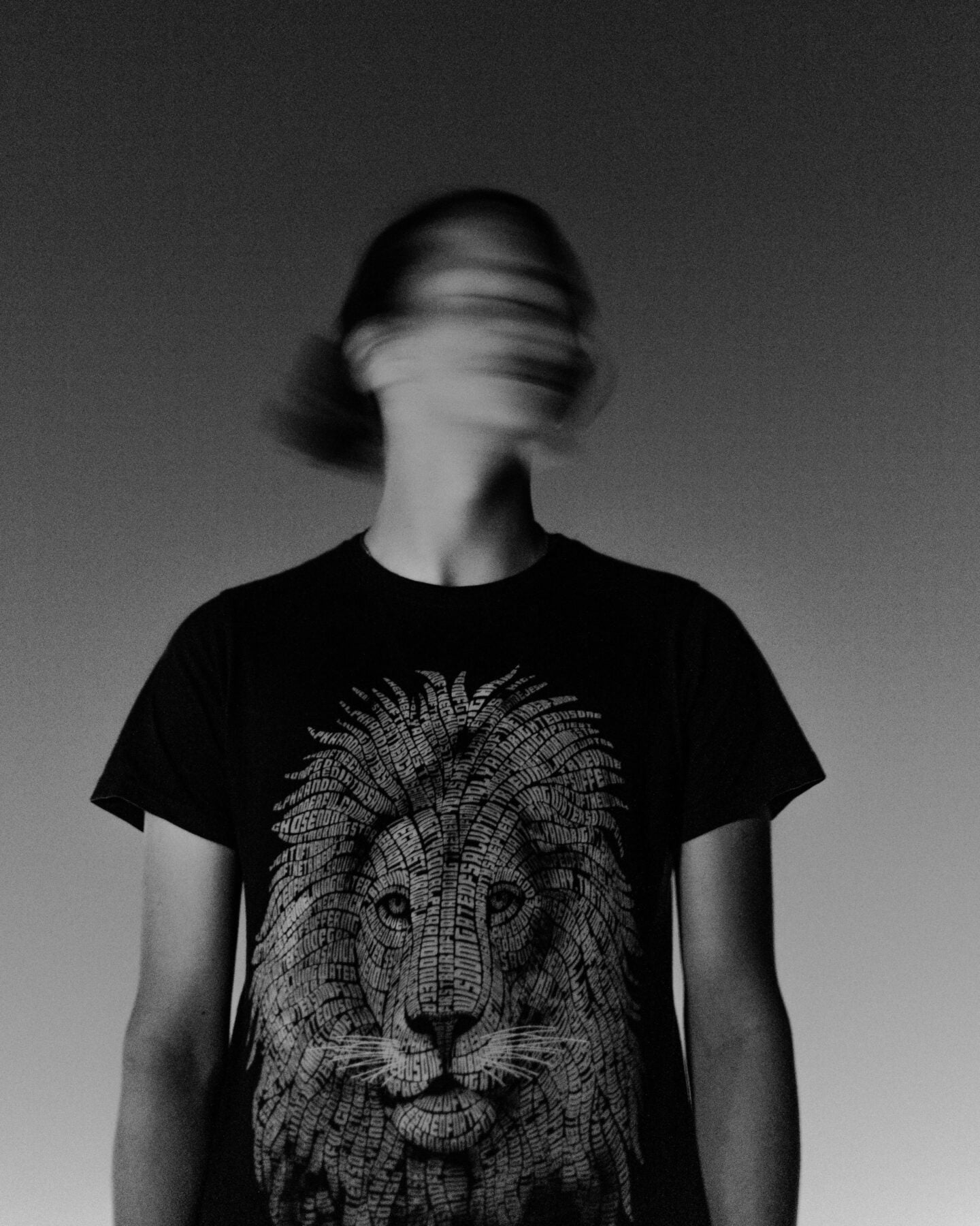 Black and white photo of a person in a black t-shirt with a detailed lion face graphic composed of layered typography; the person’s face is motion blurred.