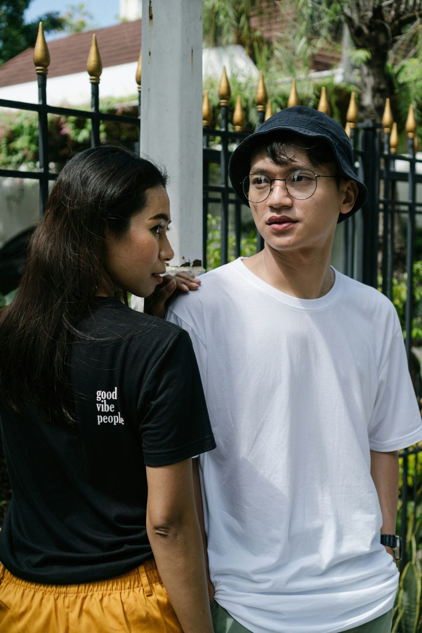 Couple standing outdoors near a black iron fence; the woman wears a black t-shirt that says "good vibe people" while the man wears a plain white t-shirt and bucket hat.