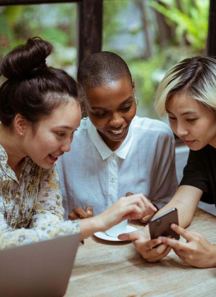 Three women smiling and engaging over a smartphone, focusing on brand monitoring and tracking online community mentions.