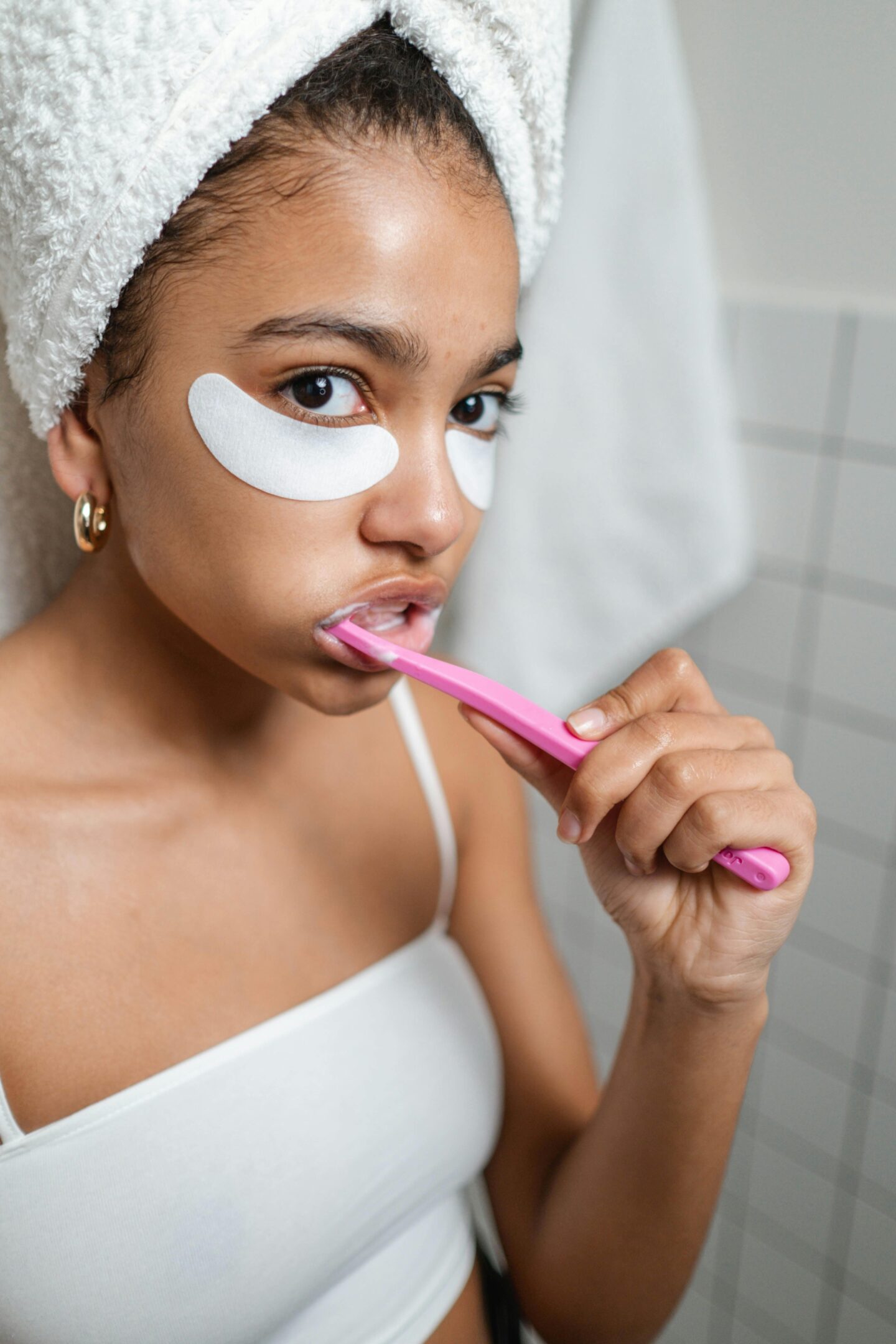 Woman with under-eye patches and a towel on her head brushing her teeth with a pink toothbrush.