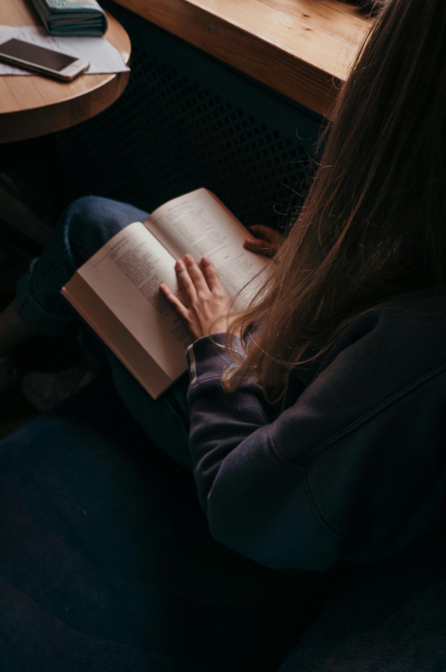 A person with long blonde hair sits by a window in a cozy setting, reading a book on their lap with a cup and phone on the table nearby.