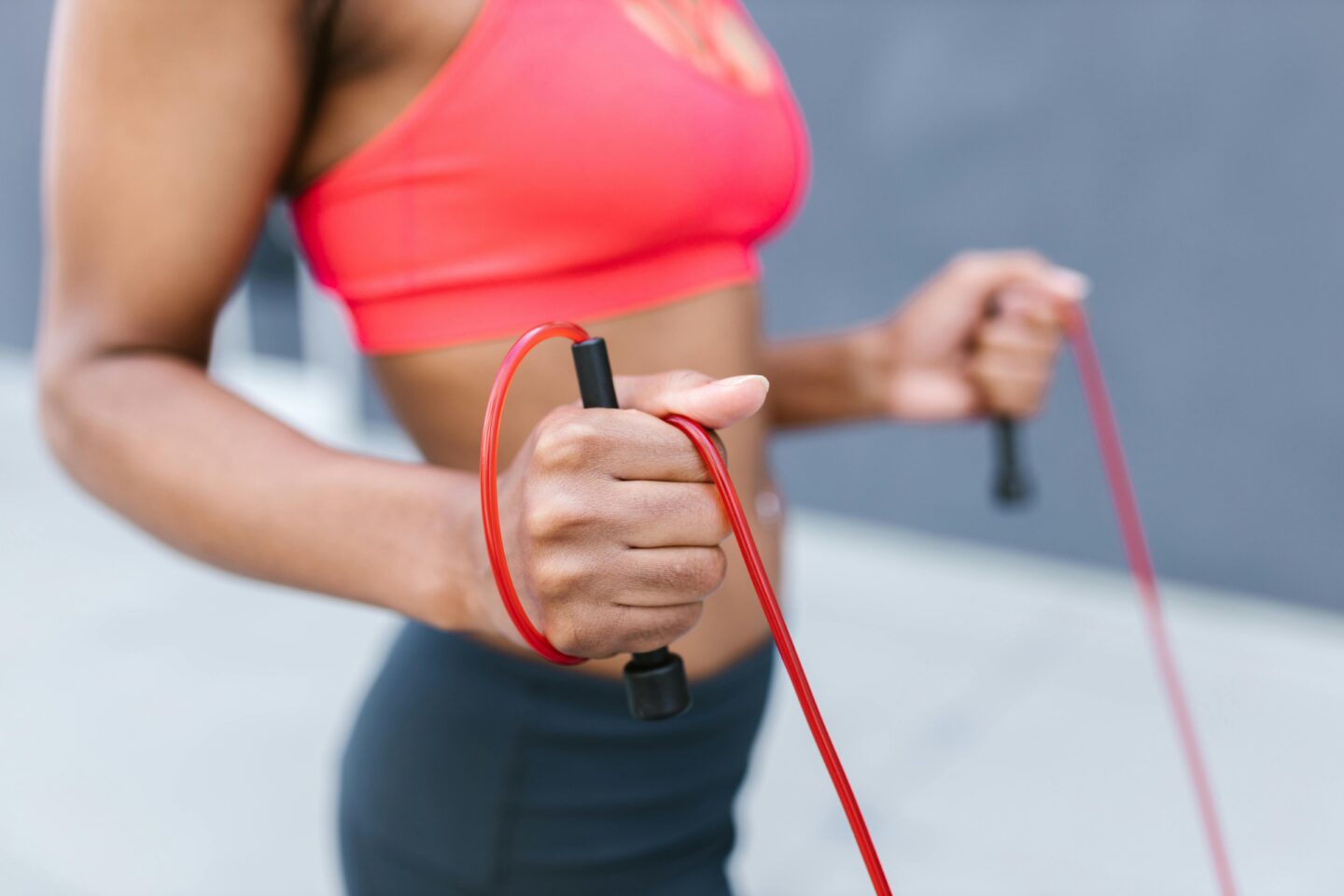 A woman in athletic wear grips a red resistance band during an outdoor workout, with focus on her toned arms and hands holding the band handles.