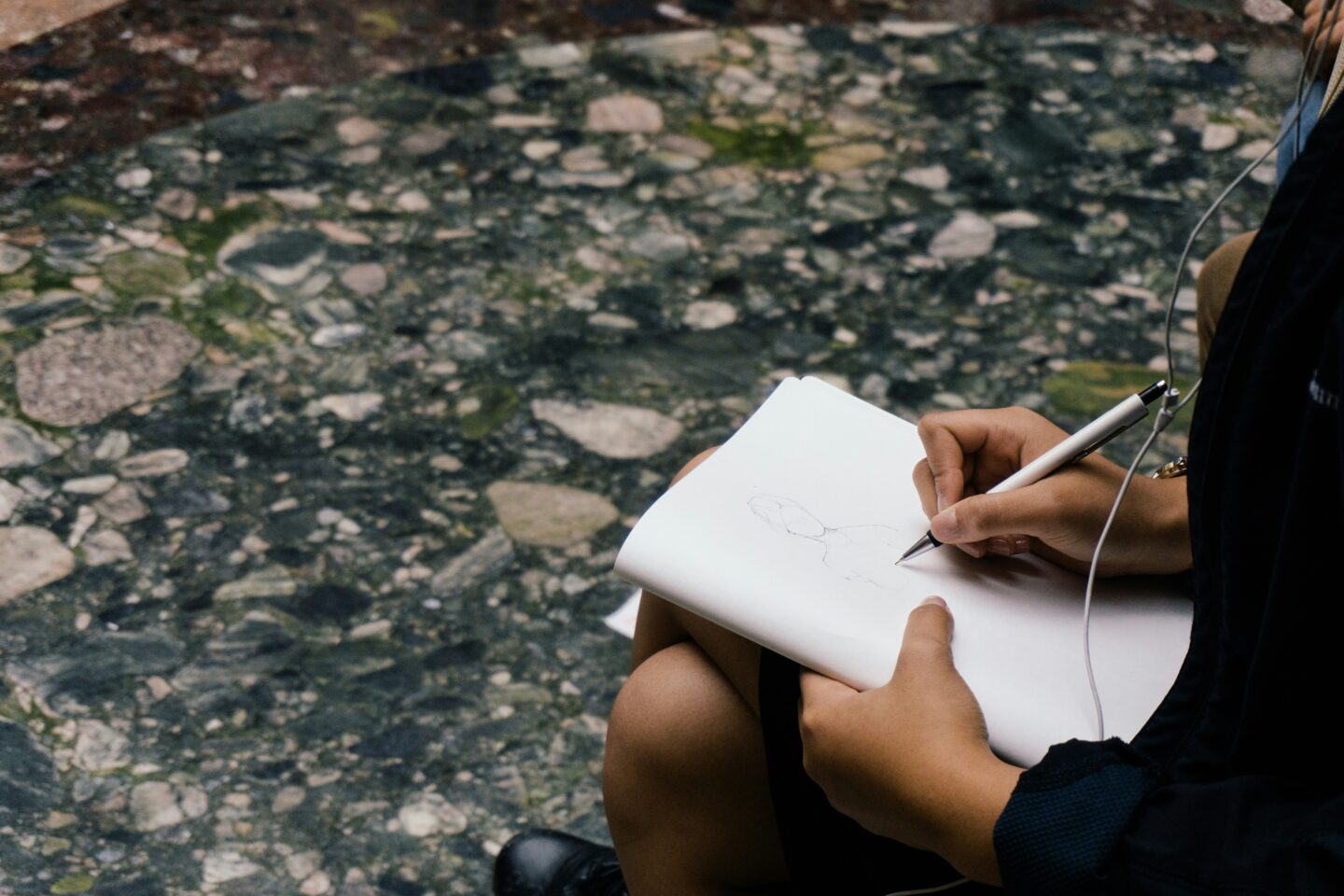 Someone sits on a stone surface, sketching a figure in a notebook while holding a pen, wearing earbuds and a dark jacket.
