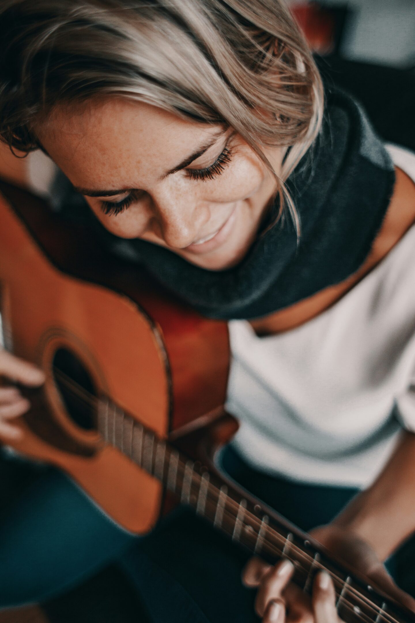 A smiling woman with short blonde hair and long lashes strums an acoustic guitar, absorbed in the moment and surrounded by a warm indoor ambiance.