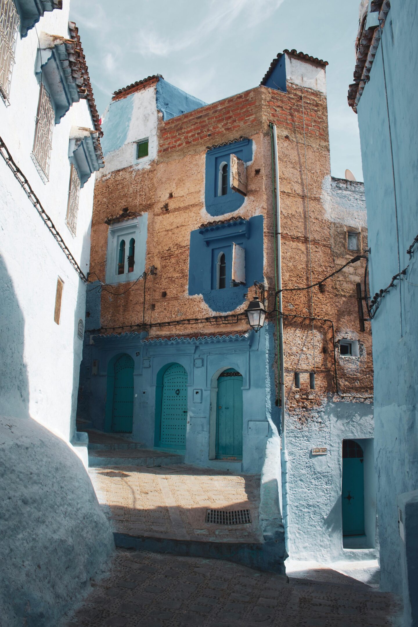 A narrow alley in Chefchaouen, Morocco, featuring buildings painted in shades of blue and earth tones with arched doorways and sunlit cobblestones.