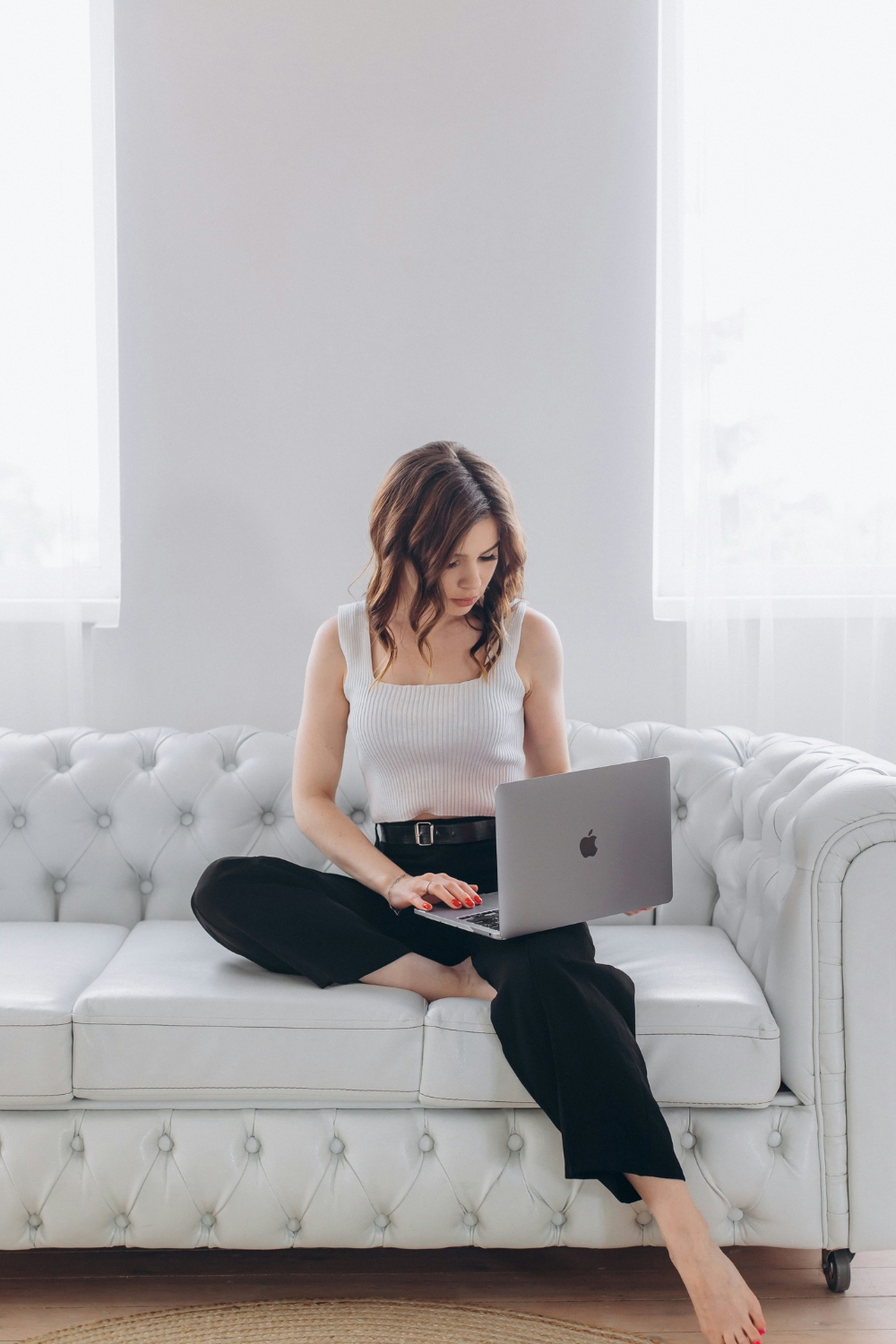Focused woman working on her business website on laptop while sitting on a white sofa in a bright modern home office.