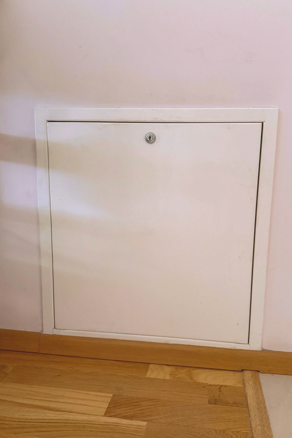 A low-mounted white access panel with a keyhole, set into a pinkish wall above a light wood floor with a matching wooden baseboard.
