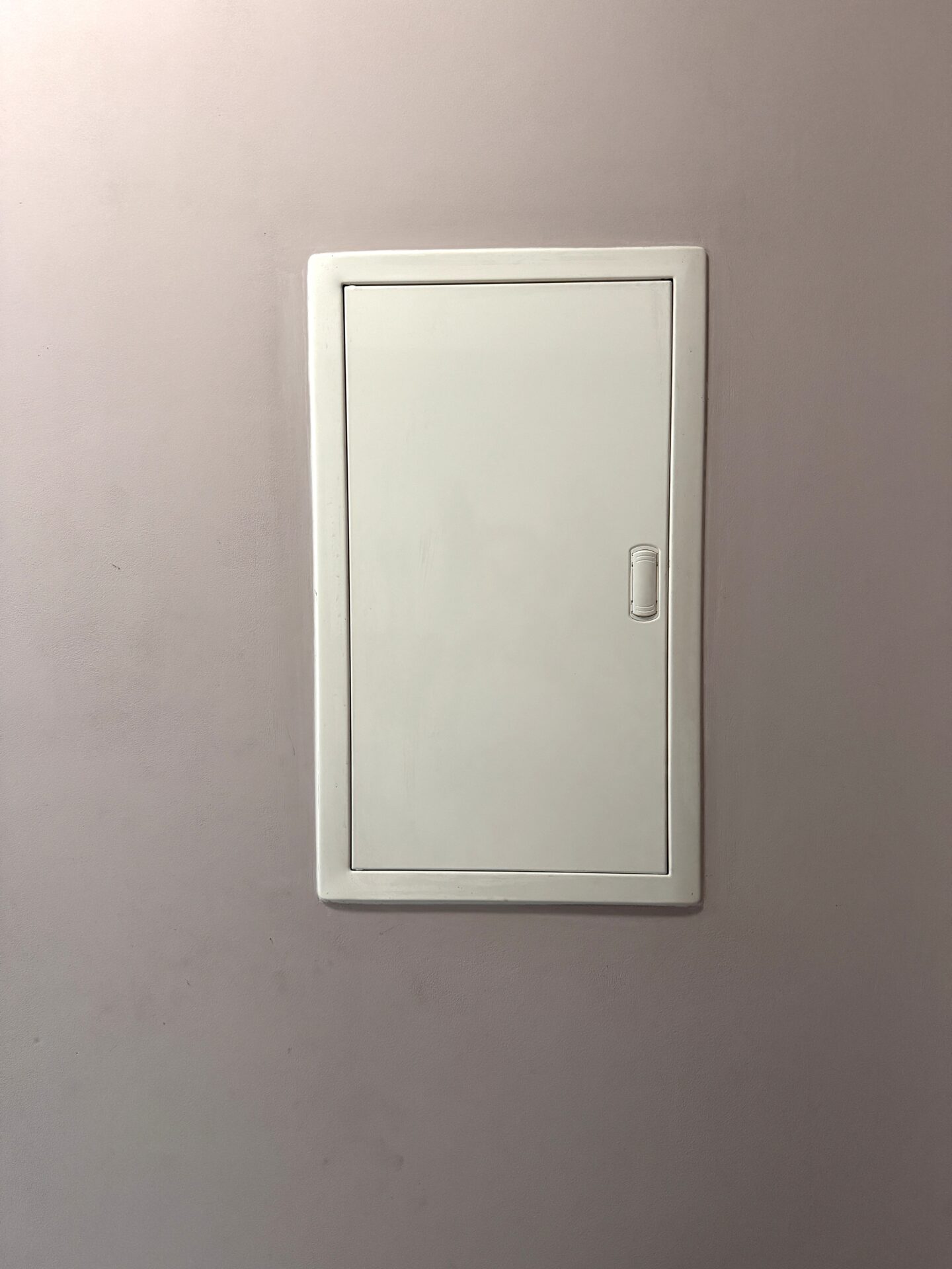 A close-up view of a vertical white metal access panel with a small rectangular recessed handle, mounted on a pale purple wall.