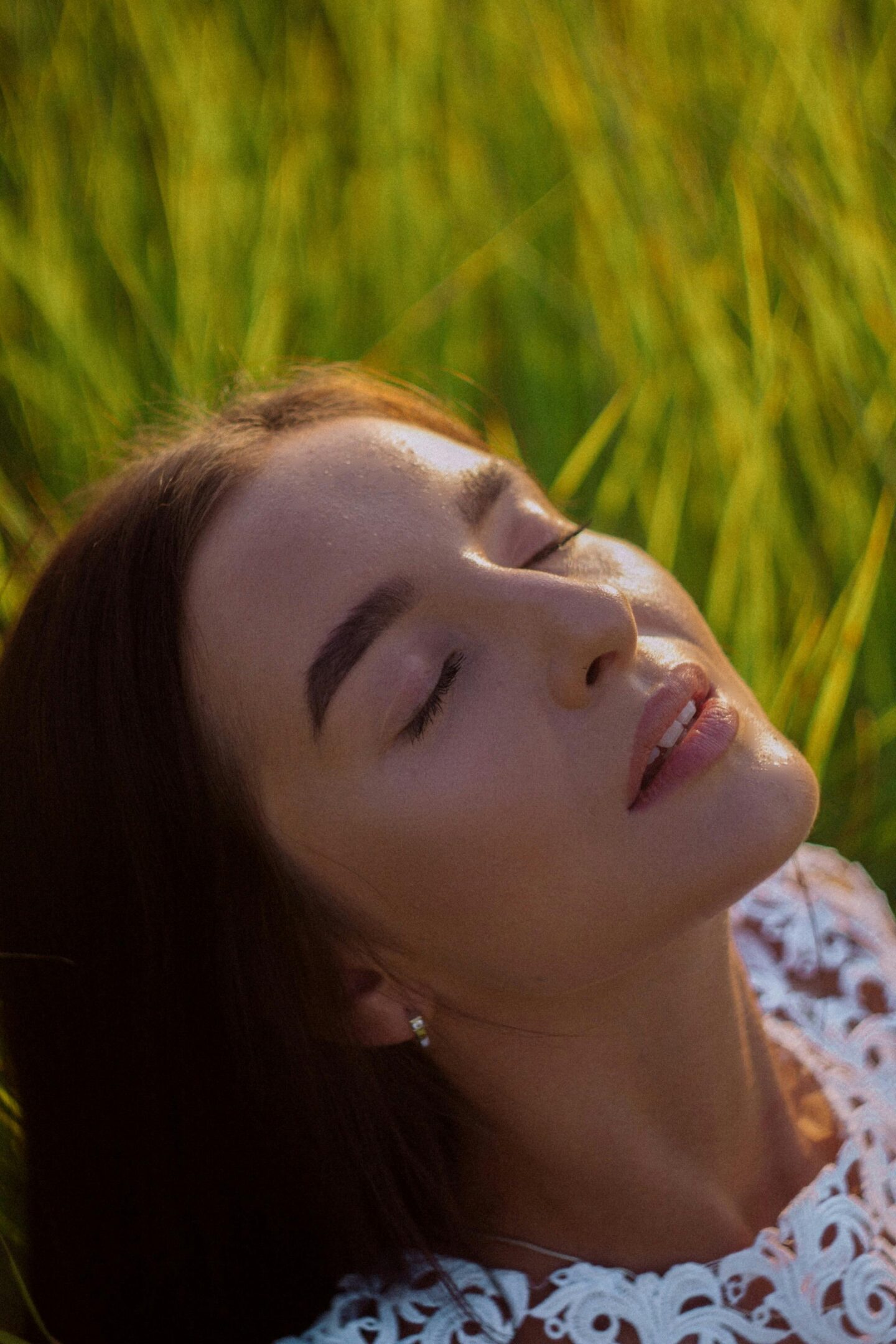 Woman with closed eyes enjoying sunlight in a grassy field, wearing a white lace top and a peaceful expression.