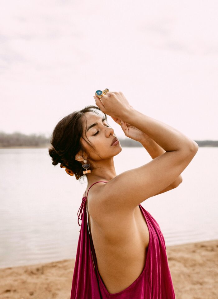 Woman standing on a sandy beach with her arms gracefully raised, eyes closed, wearing a deep pink backless dress, statement earrings, and colorful rings, with water and a soft sky in the background.