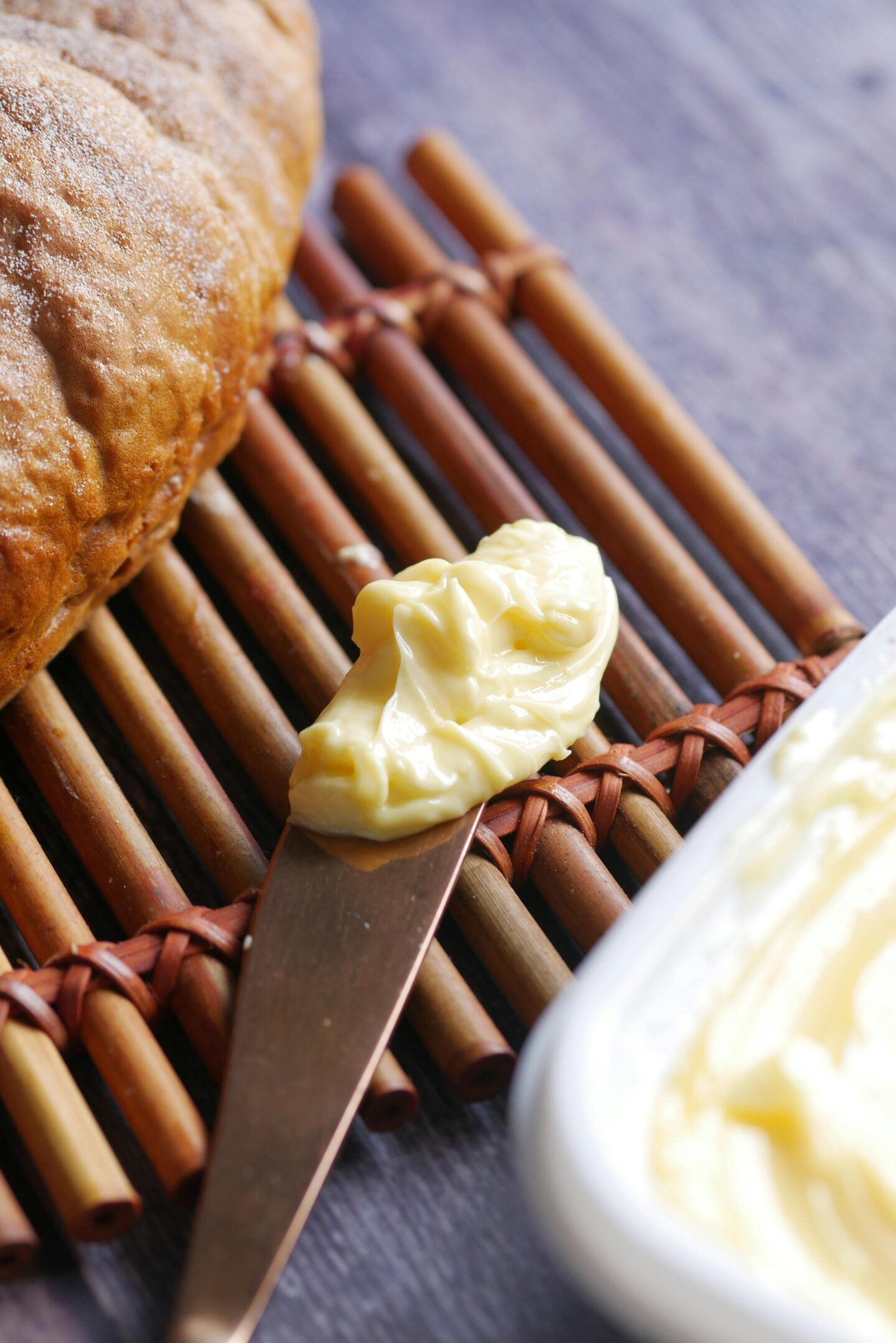 Close-up of a knife with a dollop of butter next to a crusty bread loaf on a wooden placemat.