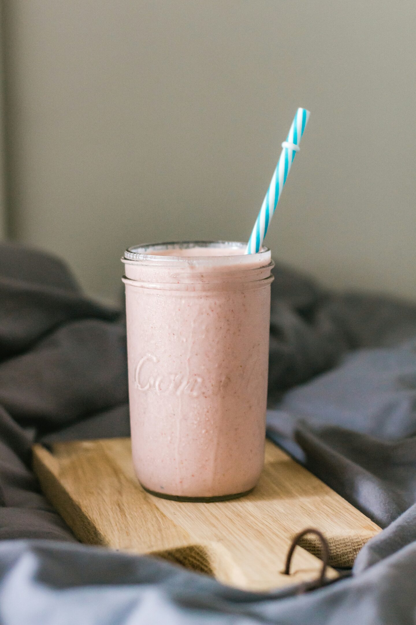 Pink smoothie served in a mason jar with a blue-striped straw, placed on a wooden board over a cozy blanket.