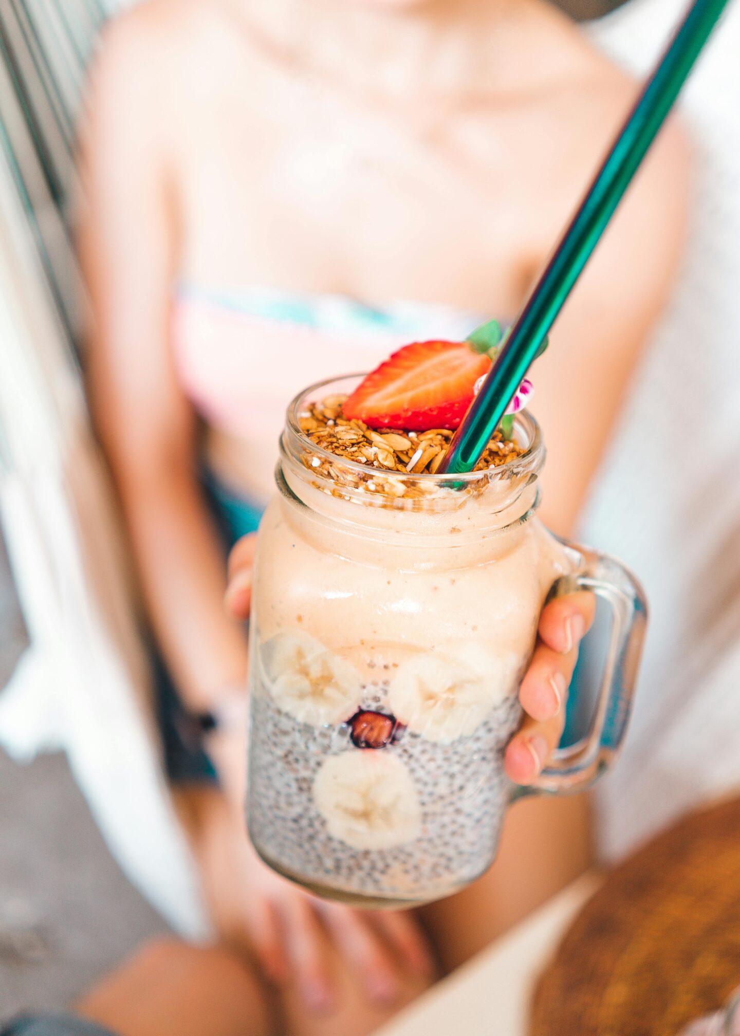 Chia pudding parfait in a mason jar with banana slices, granola, and strawberries, held by a person in a colorful swimsuit.