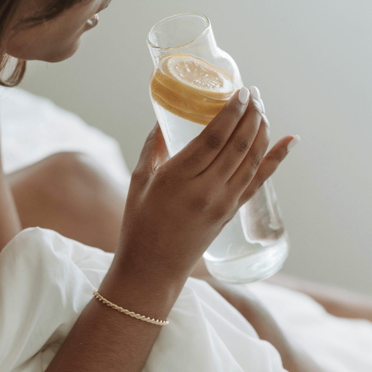 Close-up of a woman holding a glass carafe of water with lemon slices while sitting on a bed with white bedding.