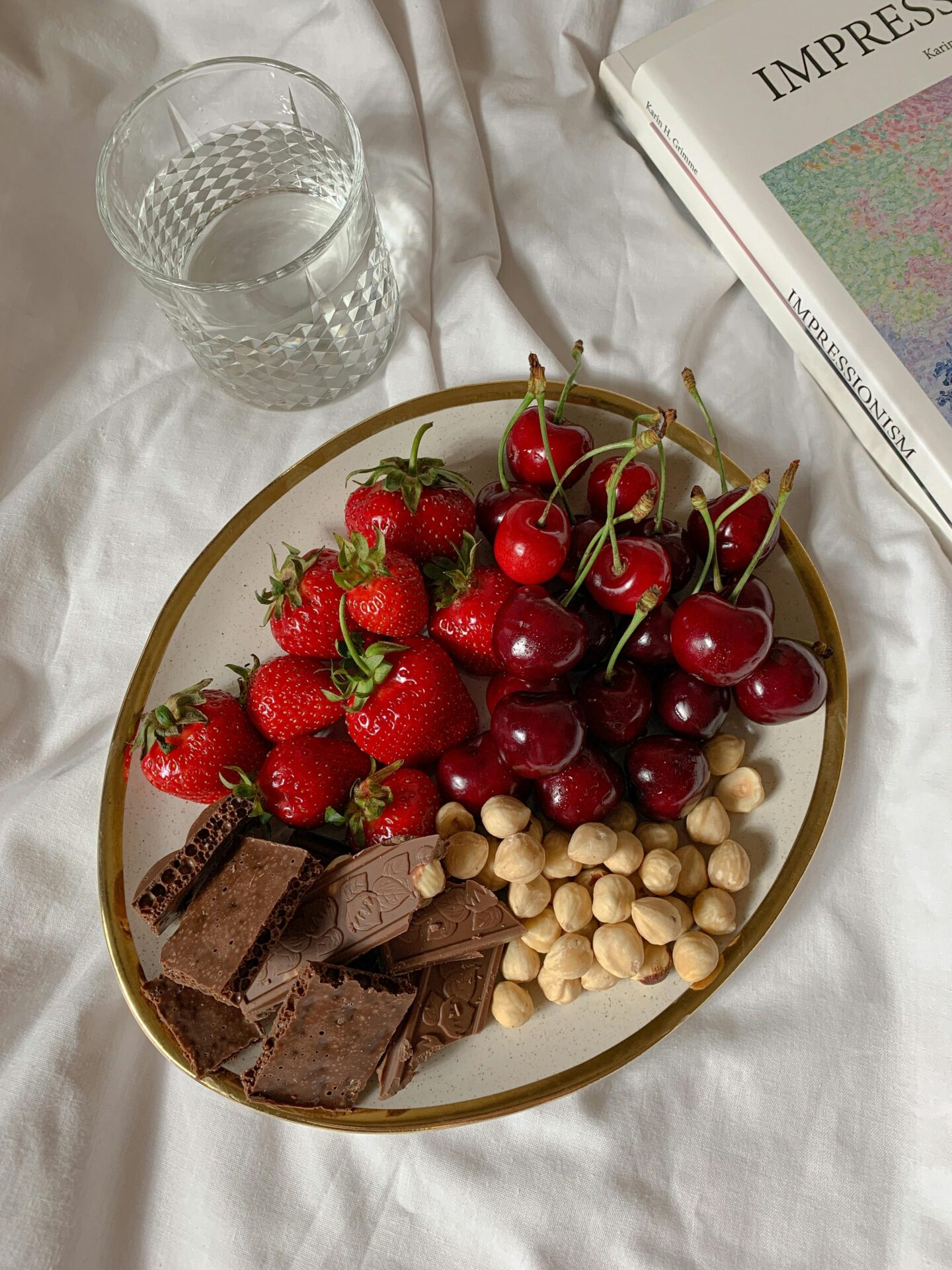 Plate of fresh strawberries, cherries, hazelnuts, and chocolate pieces on a bed with a glass of water and an Impressionist art book nearby.