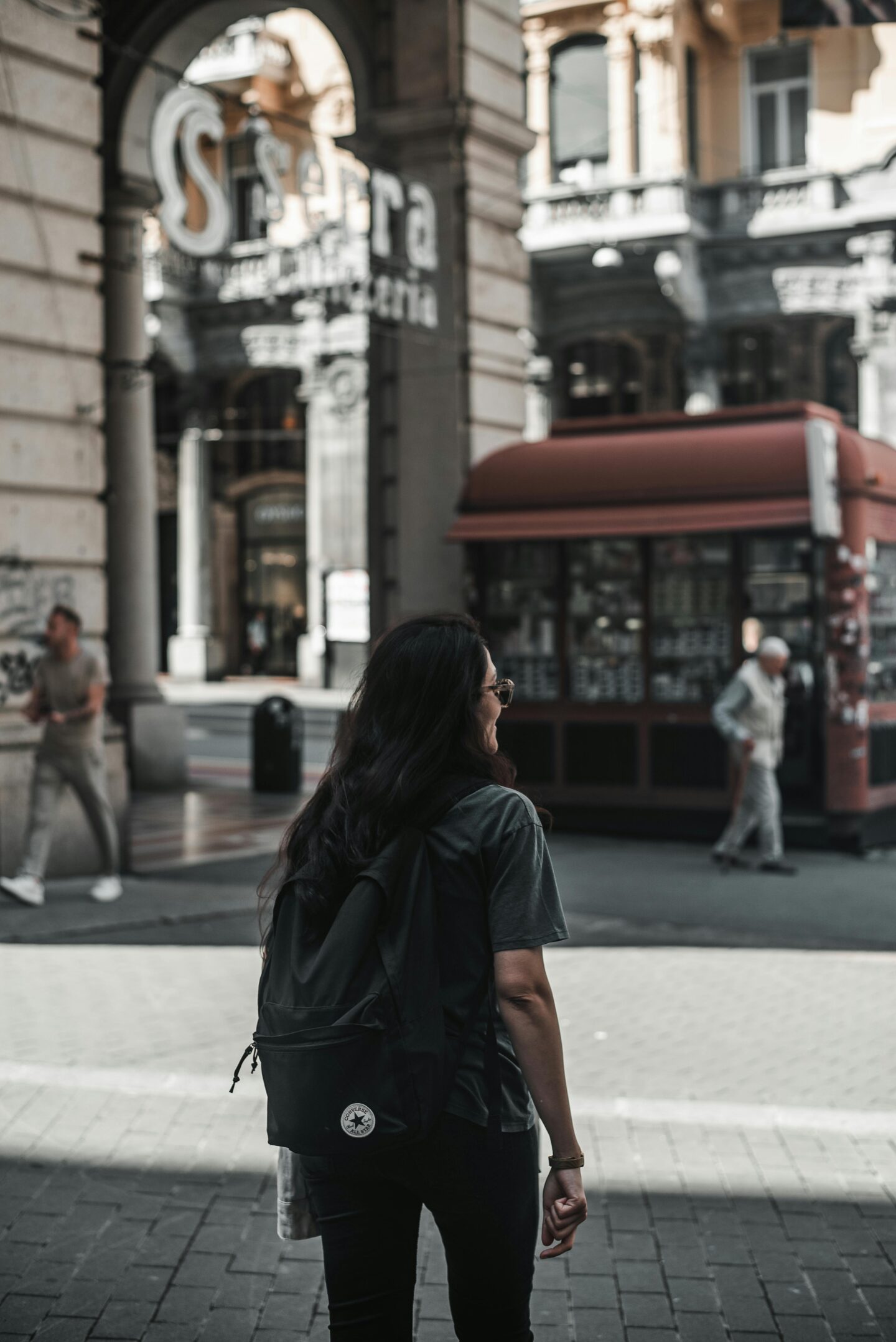 Woman walking with a backpack through a historic European shopping arcade in warm afternoon light.
