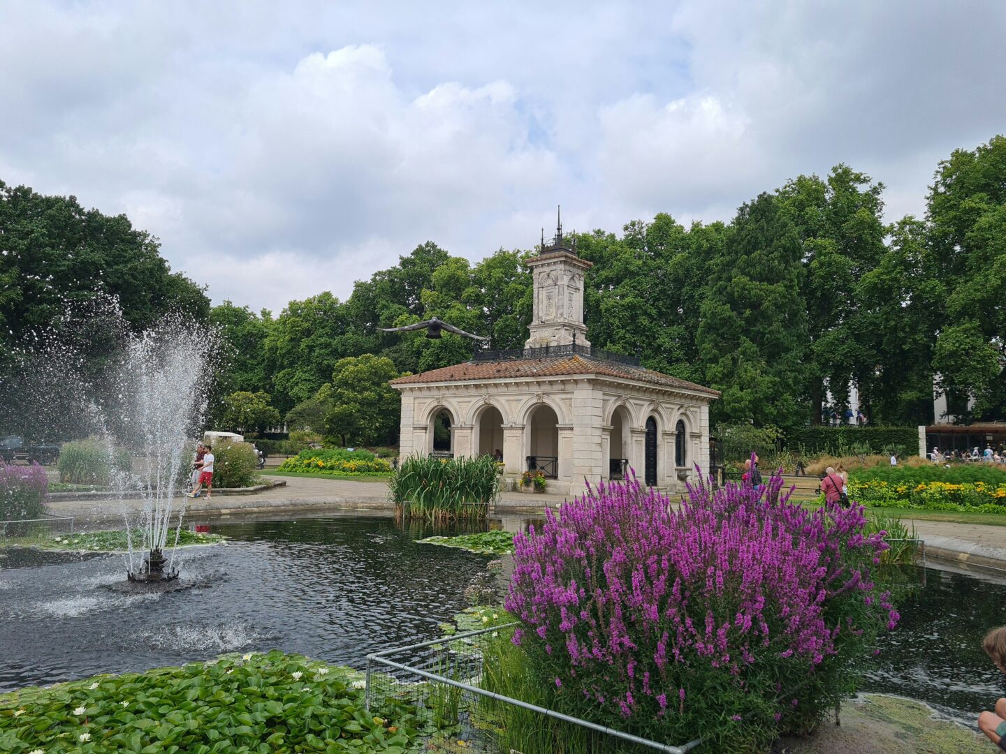 Fountain and blooming purple flowers in the Italian Gardens of Hyde Park with a classical stone building in the background.