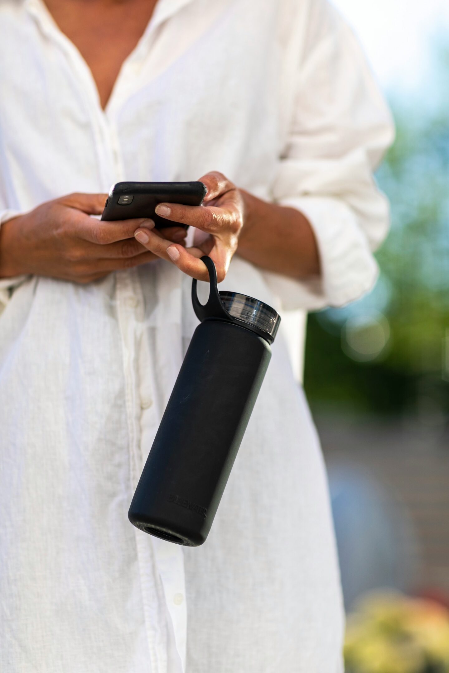 Person wearing a white linen shirt while holding a smartphone and reusable water bottle outdoors in summer light.