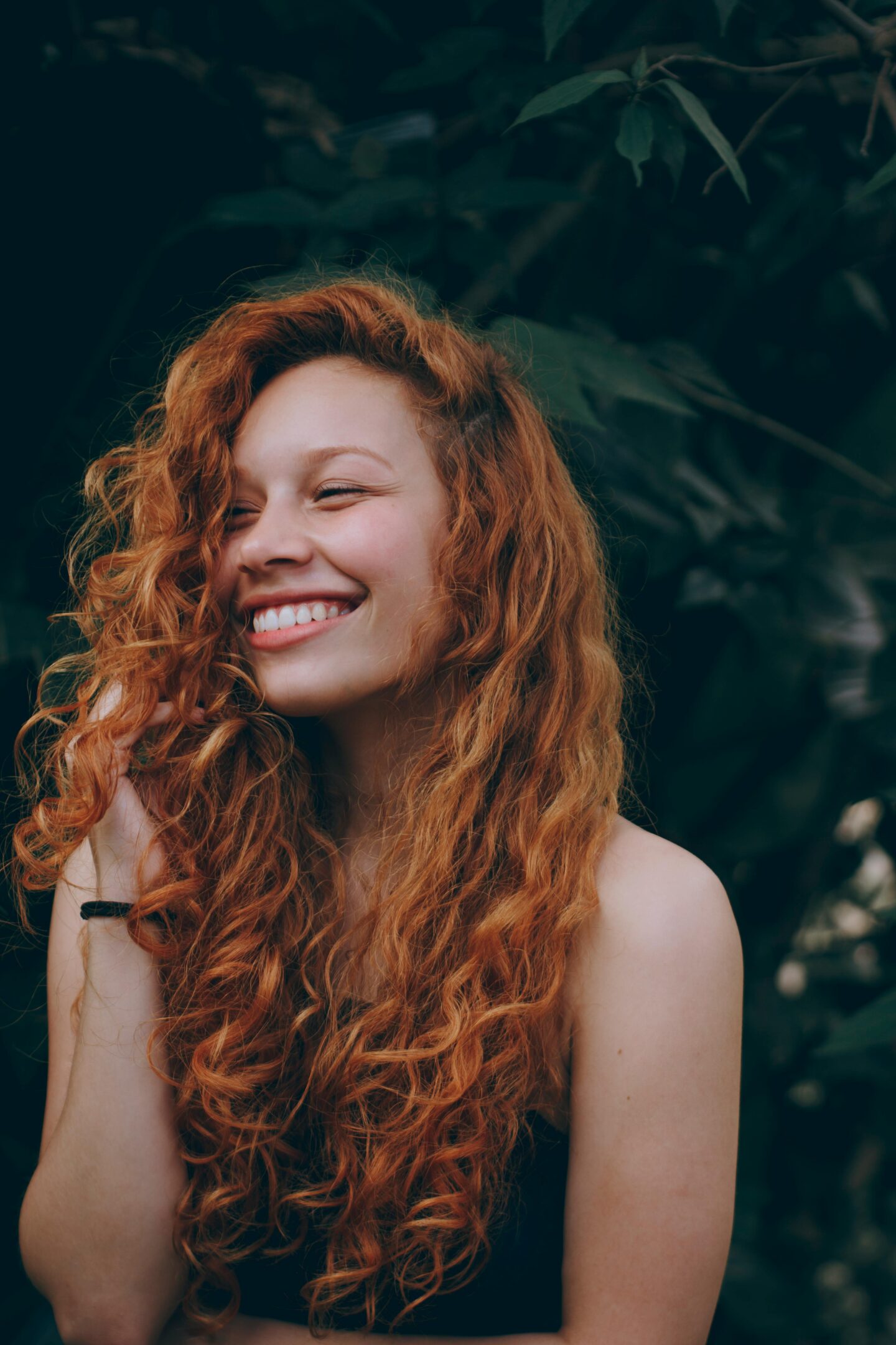 A woman with long curly red hair smiles joyfully, partially covering her face with her hair, with a dark green leafy background.