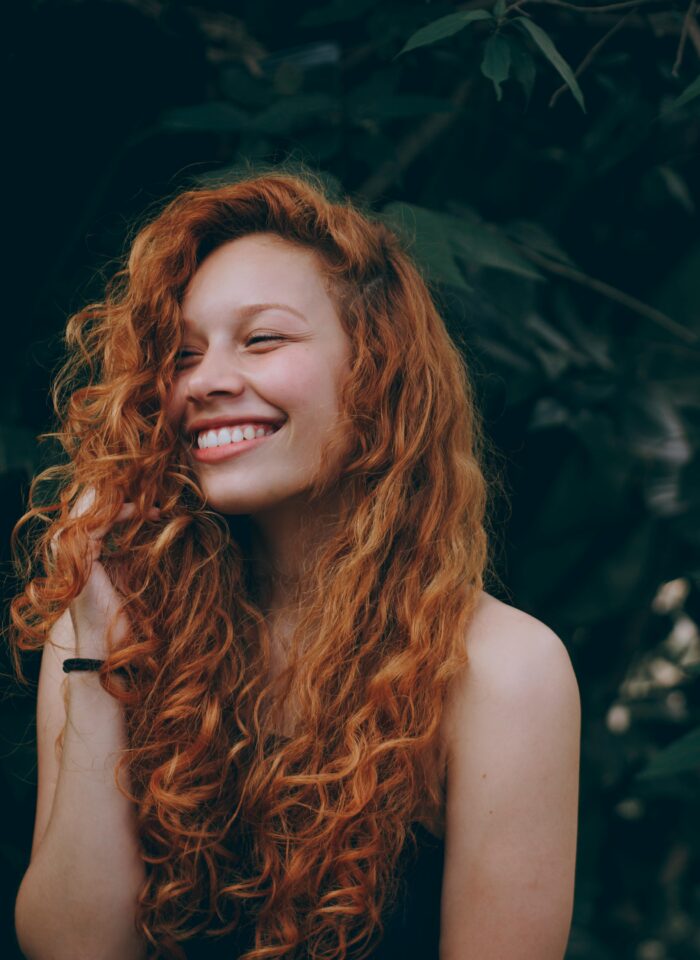 A woman with long curly red hair smiles joyfully, partially covering her face with her hair, with a dark green leafy background.
