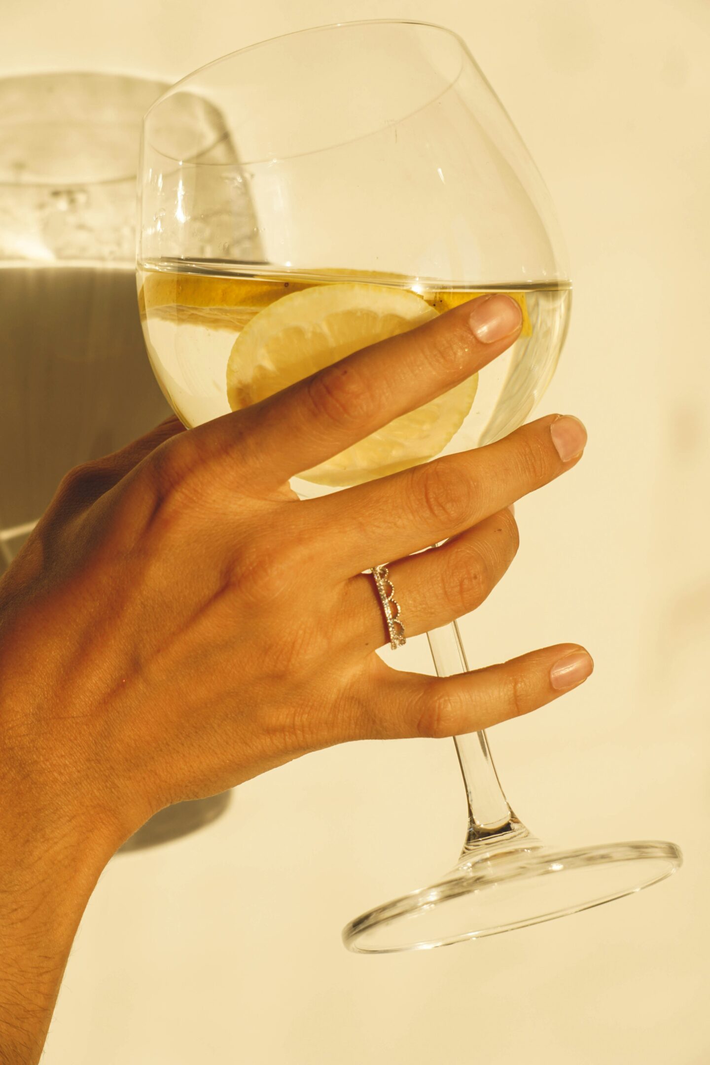 A hand holding a wine glass fillied with water with lemon slices, wearing a delicate ring with round gemstones, lit by warm sunlight.