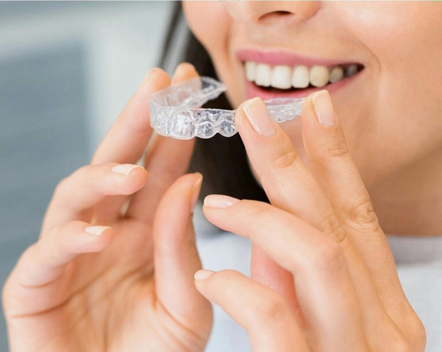 A close-up of a smiling woman holding a clear dental aligner between her fingers, preparing to place it in her mouth.