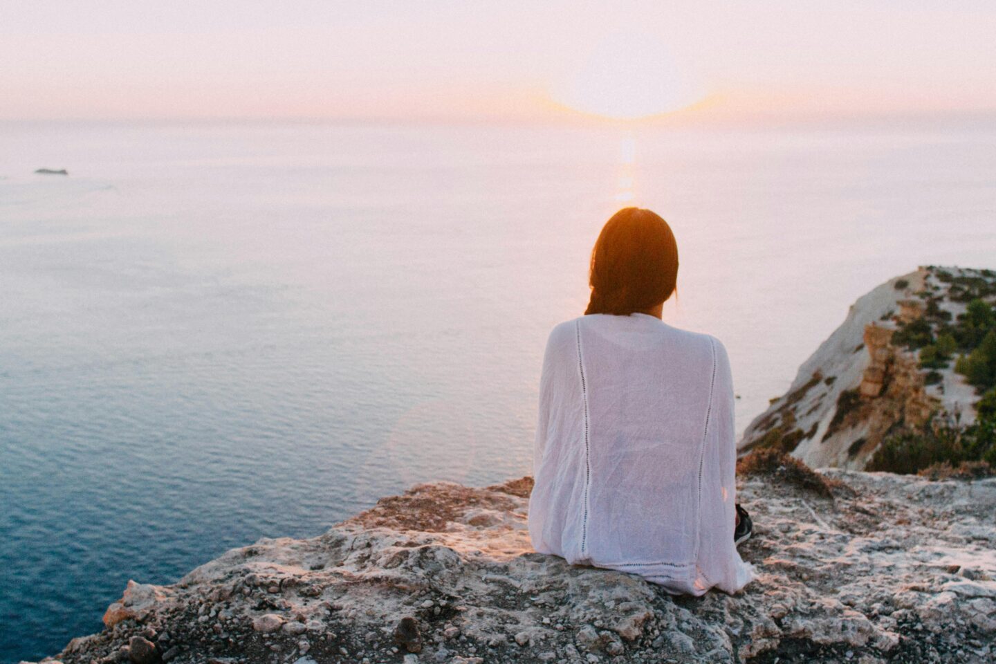 Woman in a white top sitting alone on a cliff at sunset, reflecting on life and embracing solitude for emotional healing and clarity.