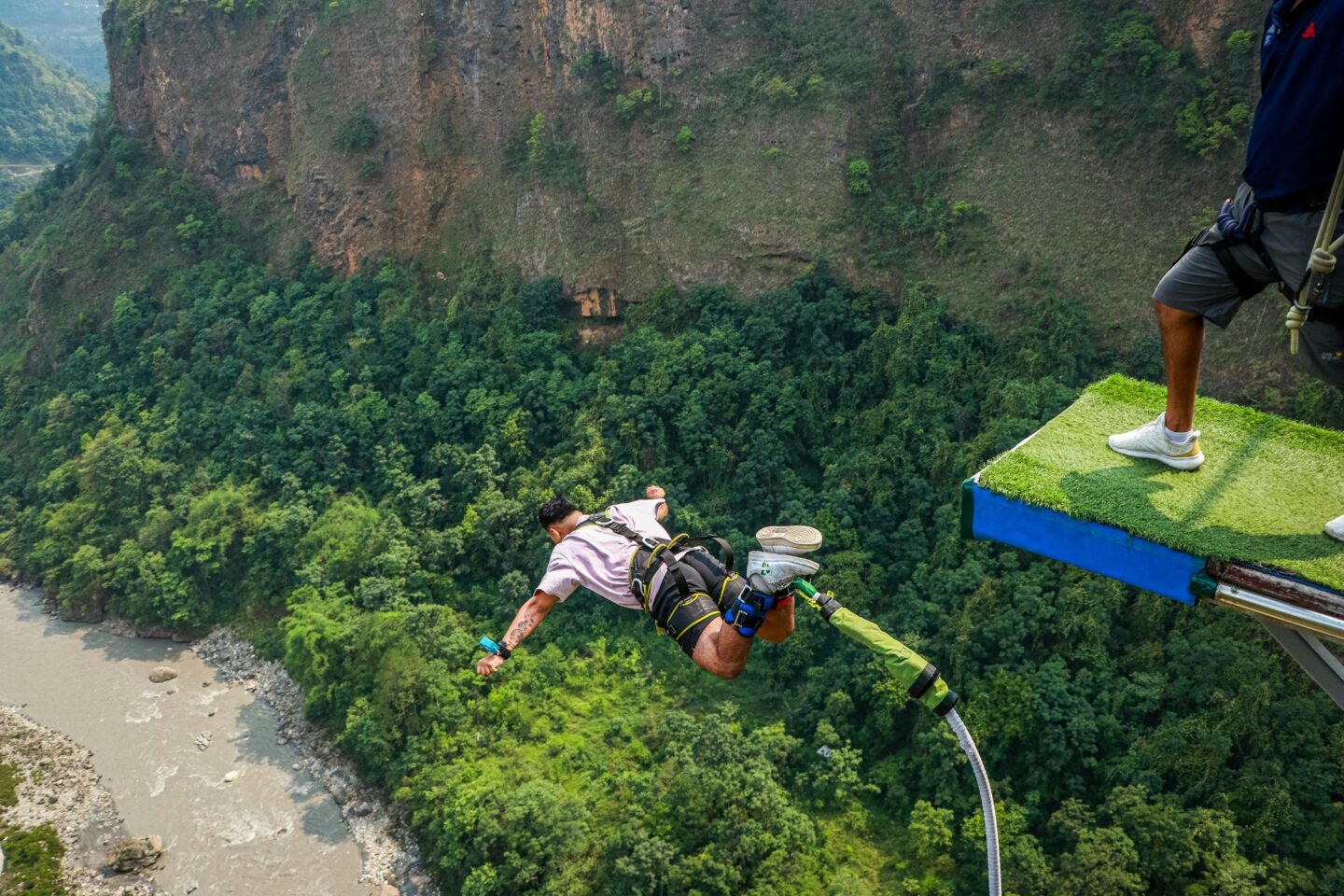 Person in a harness mid-air during a bungee jump off a high platform above a lush green valley and river.