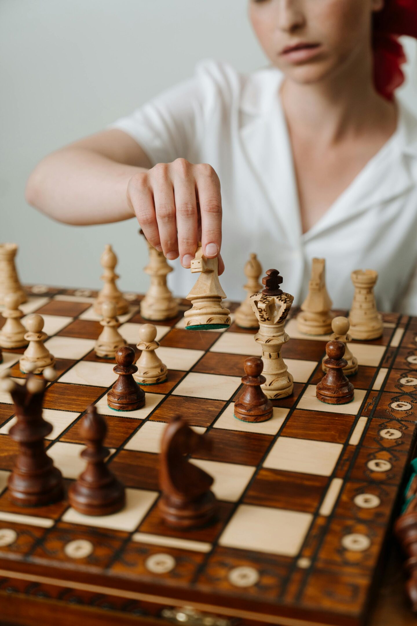Woman in a white shirt playing chess, holding a knight piece over a wooden chessboard with ornate detailing.