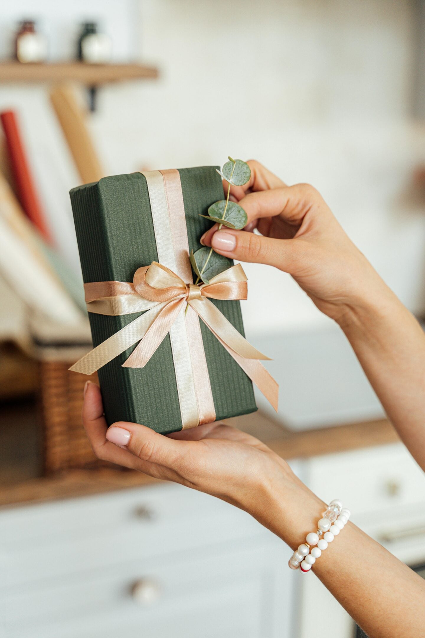 Close-up of hands holding a green gift box tied with a satin ribbon and decorated with eucalyptus leaves.