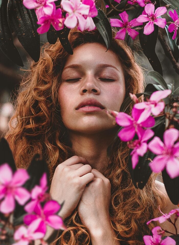 Woman with curly hair surrounded by pink flowers, practicing mindfulness outdoors.