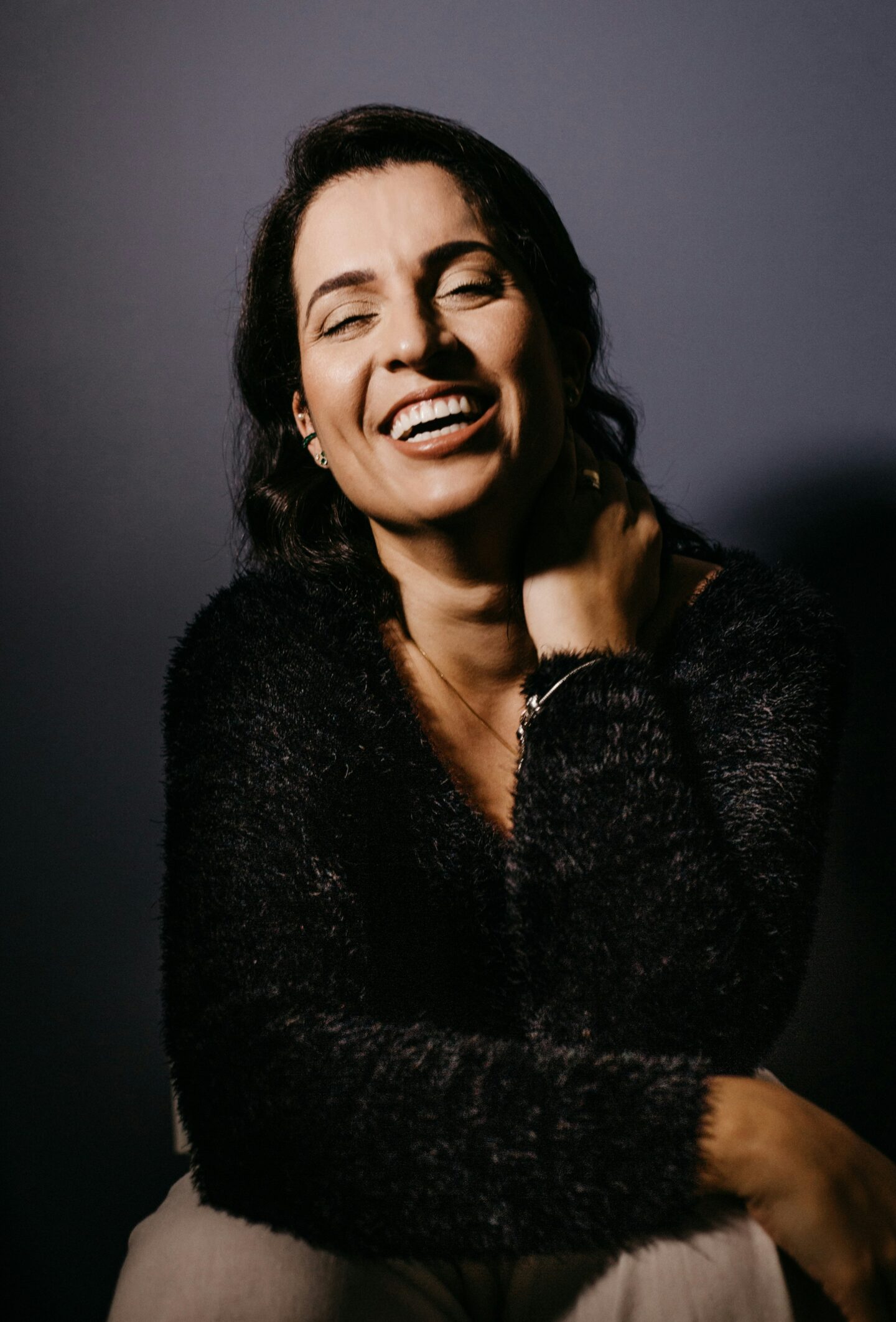 A smiling woman with dark hair sitting in front of a dark background, wearing a fuzzy black sweater and holding her neck casually with one hand.