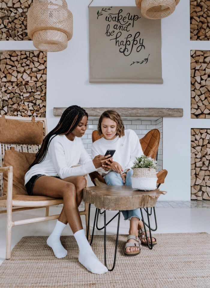 Two women sitting indoors in cozy chairs, looking at a smartphone together, with a wooden coffee table and stacked firewood in the background.