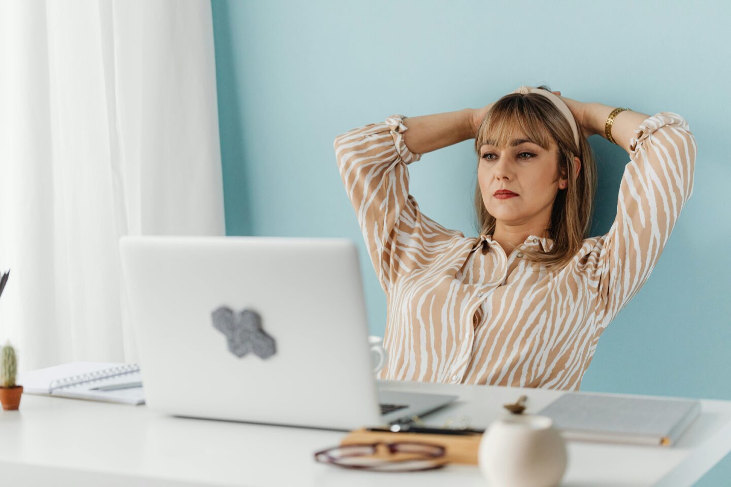 A woman sitting at a desk with a laptop, leaning back with her hands behind her head, wearing a beige and white striped blouse and looking thoughtful.