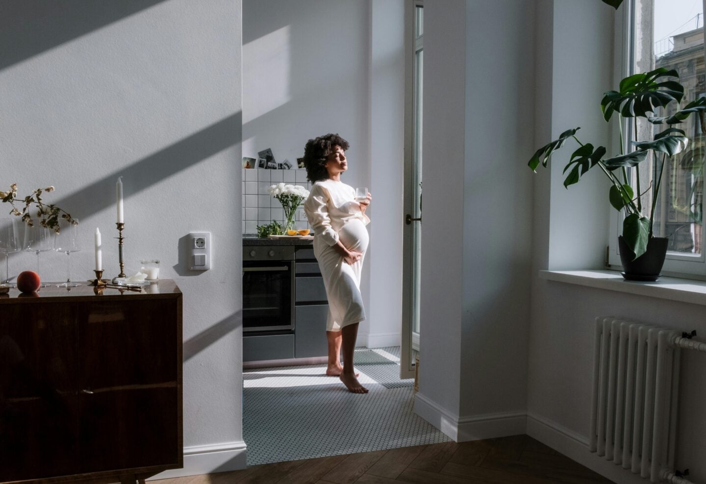 Pregnant woman standing in a sunlit modern kitchen holding a glass of water, surrounded by plants.