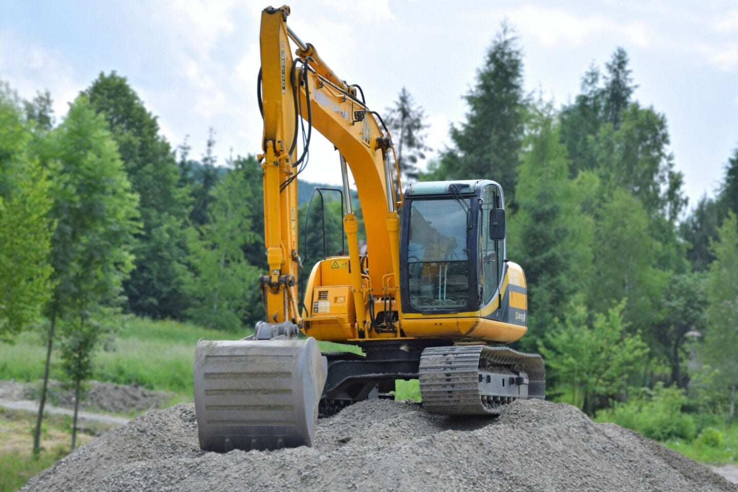 Large yellow excavator on a mound of gravel at a construction site with trees in the background.