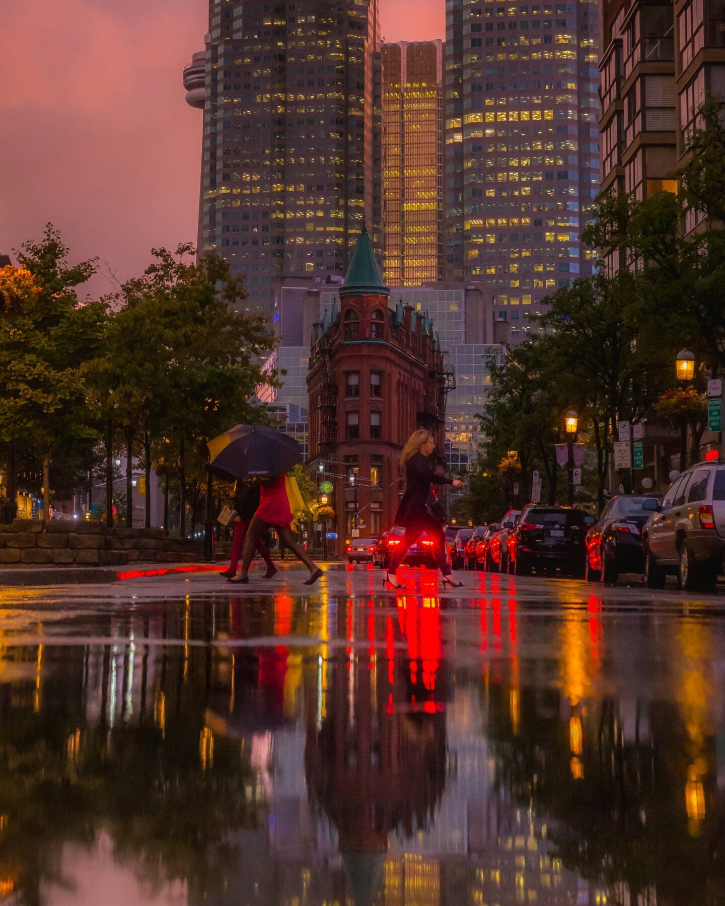 Rainy Toronto city street at dusk with reflections of lights on wet pavement and pedestrians with umbrellas.