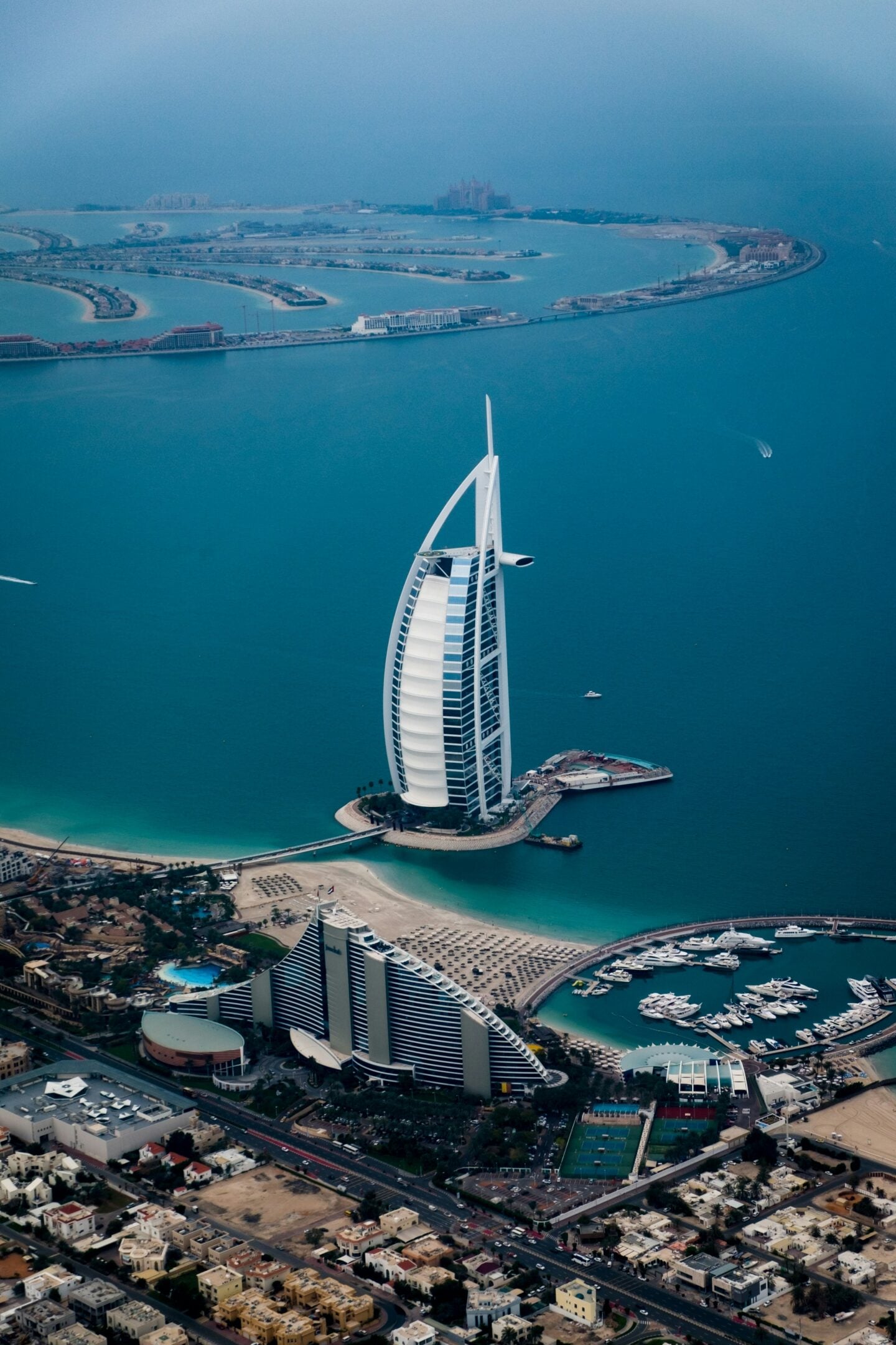 Aerial view of the Burj Al Arab hotel on its own island with the Palm Jumeirah and Dubai coastline in the background.