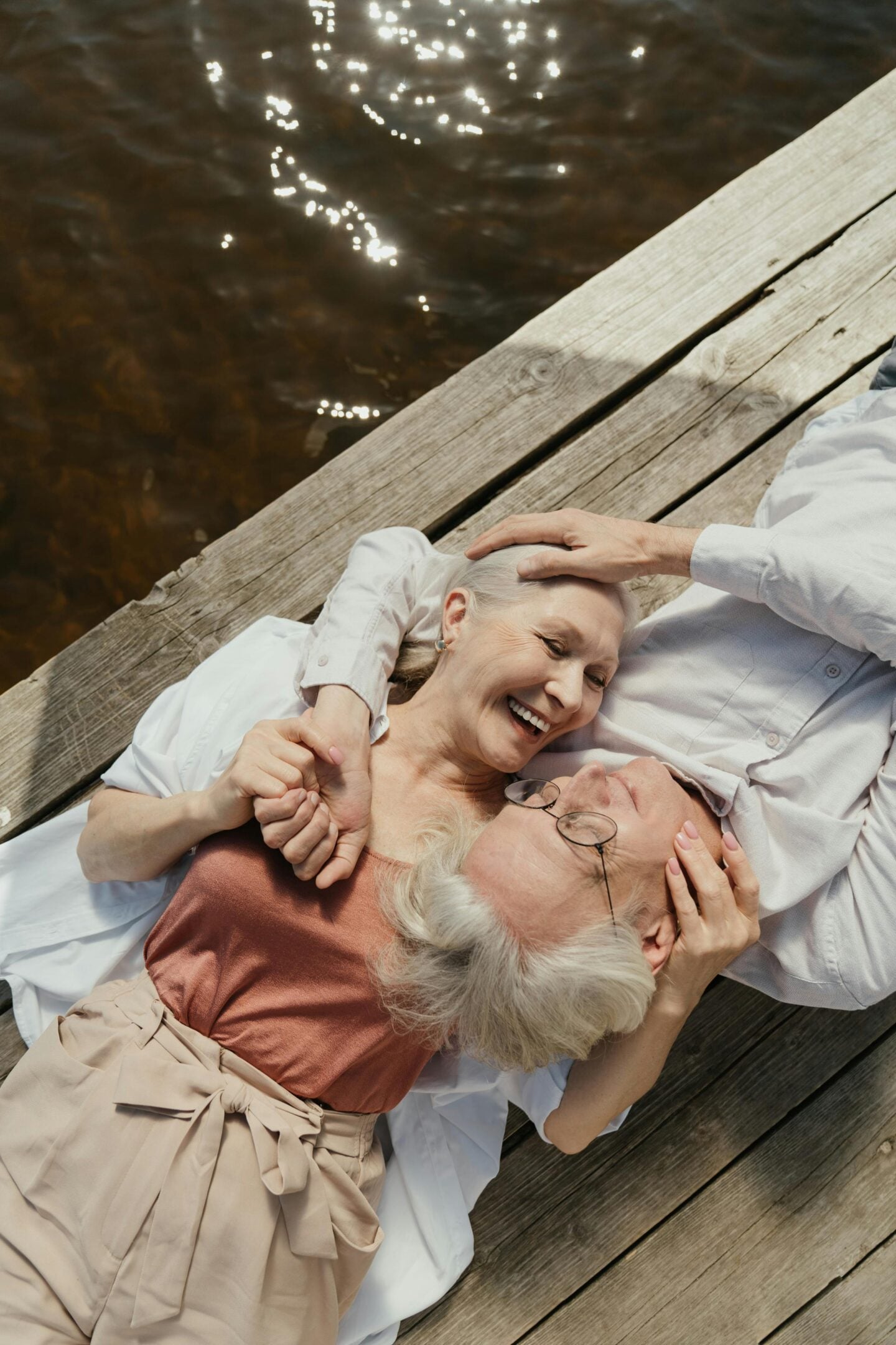 Elderly couple lying side by side on a wooden dock near water, holding each other and laughing in a moment of affection.