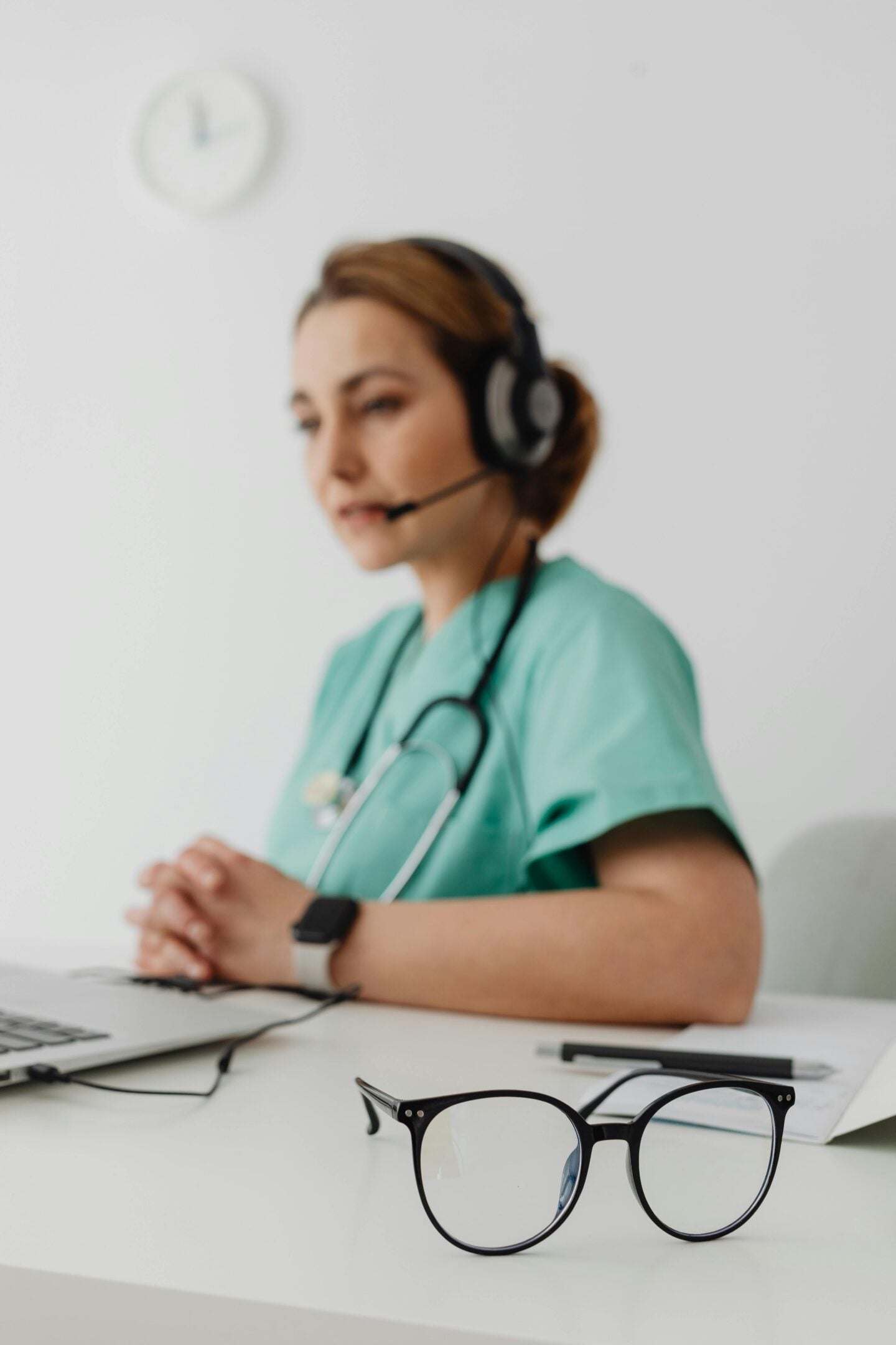 Close-up of a pair of eyeglasses on a desk with a female healthcare professional wearing a headset and scrubs blurred in the background.