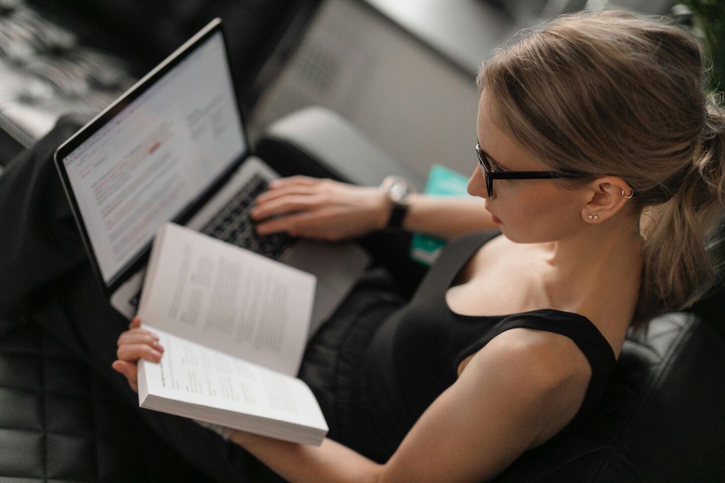 Young woman wearing glasses reading a book and working on a laptop.