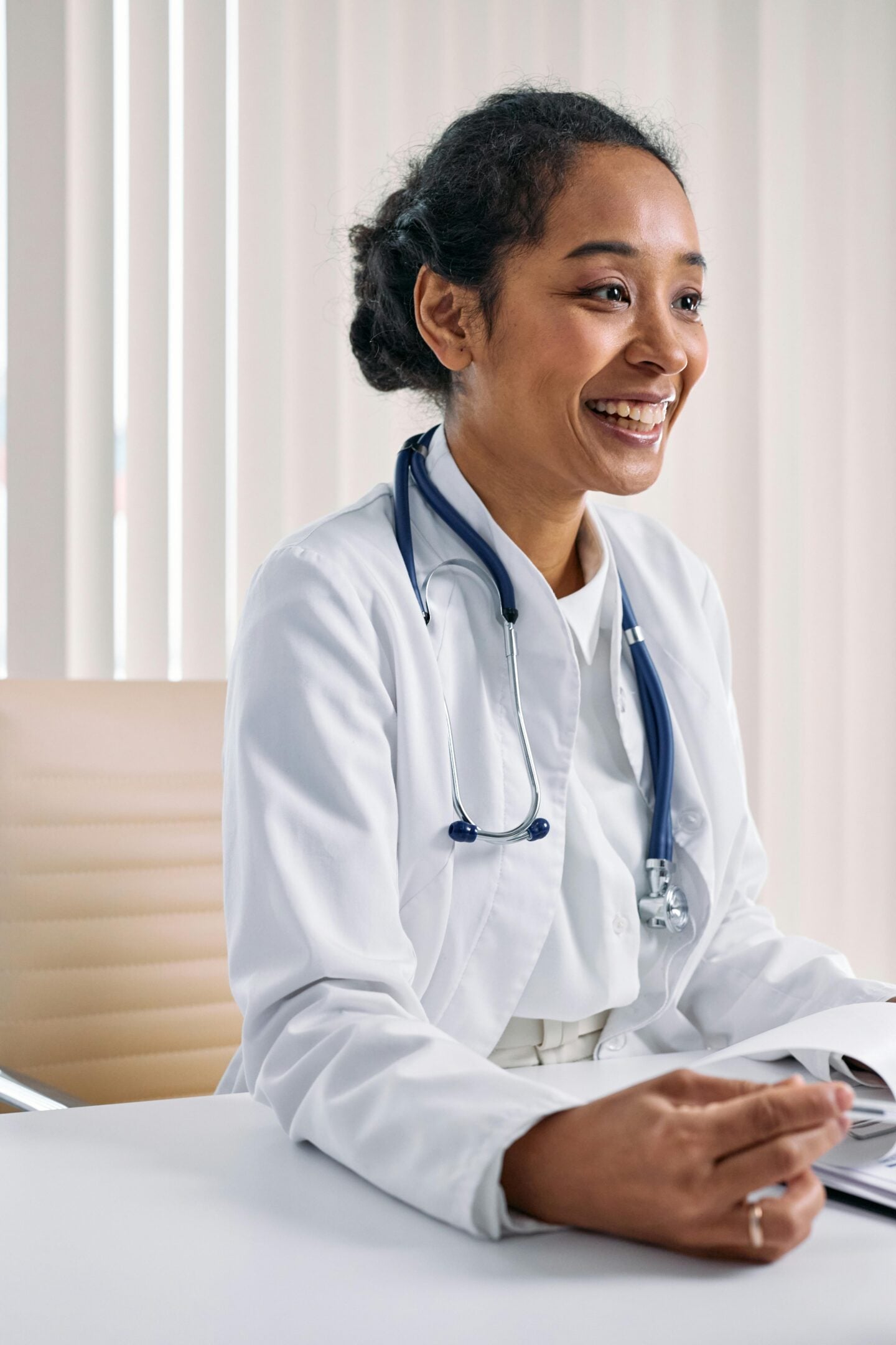 Cheerful female doctor wearing a white coat and stethoscope sitting at a desk during a consultation.